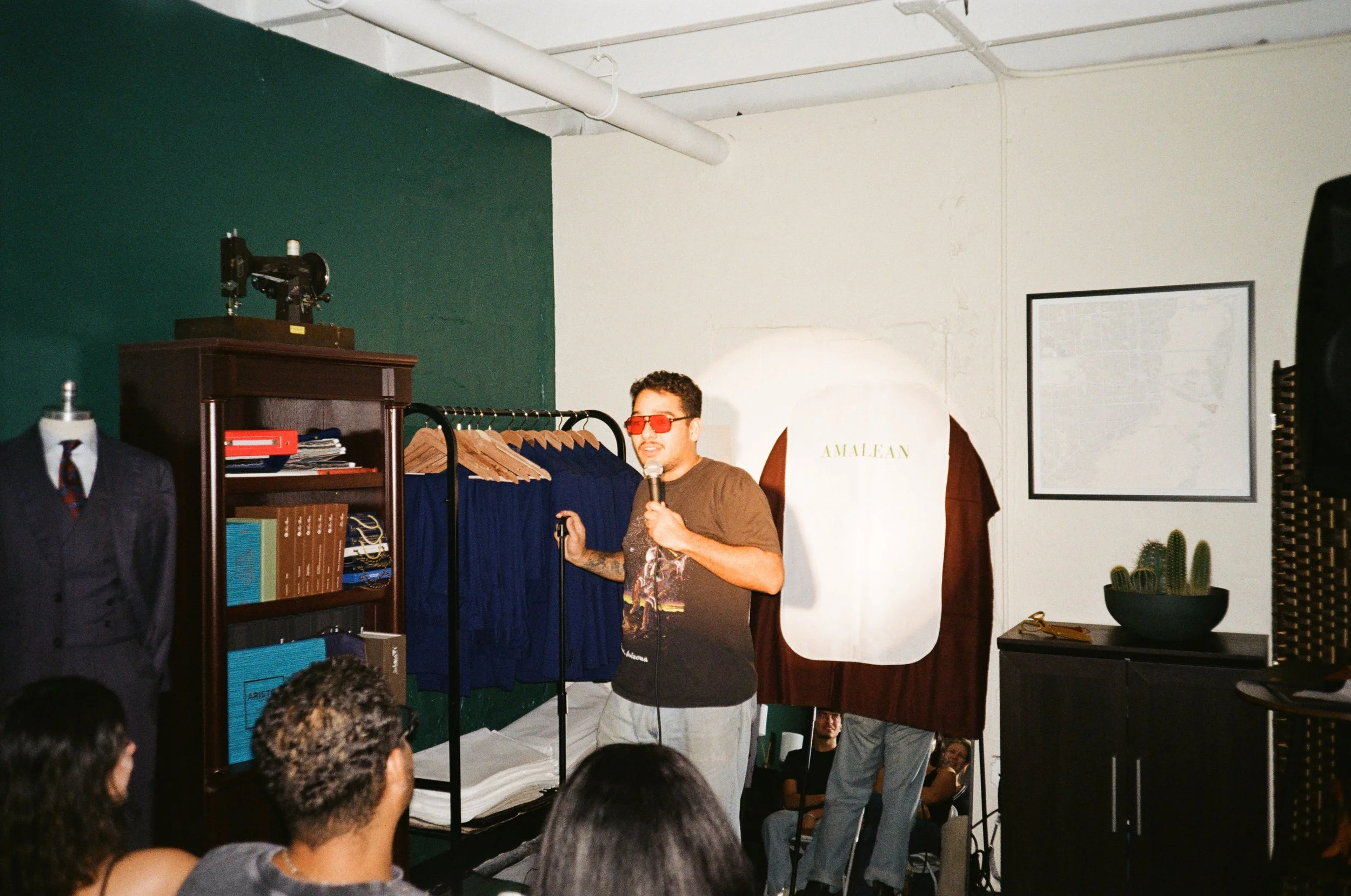 A man with dark curly hair wearing red glasses, a black graphic t-shirt, and gray pants holding a microphone, standing next to a clothing rack with blue garments in a room with white and dark green walls. A mannequin with a suit, a bookshelf with boo