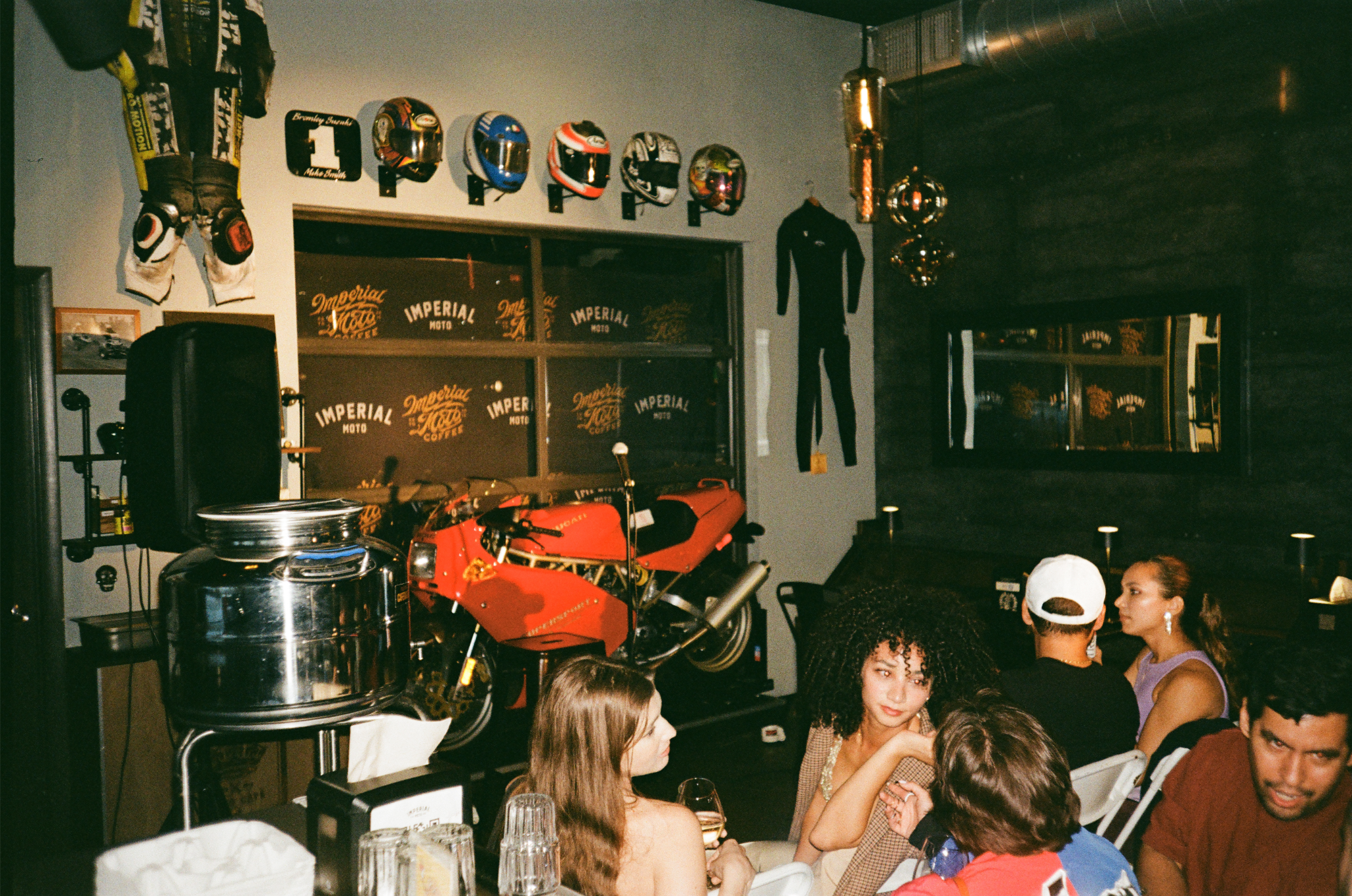 A group of people sitting at a table in a motorcycle-themed bar or restaurant. The wall behind them displays racing helmets, a racing suit, and a motorcycle, along with vintage signs that read Imperial Moto Coffee. There are other decorative elements