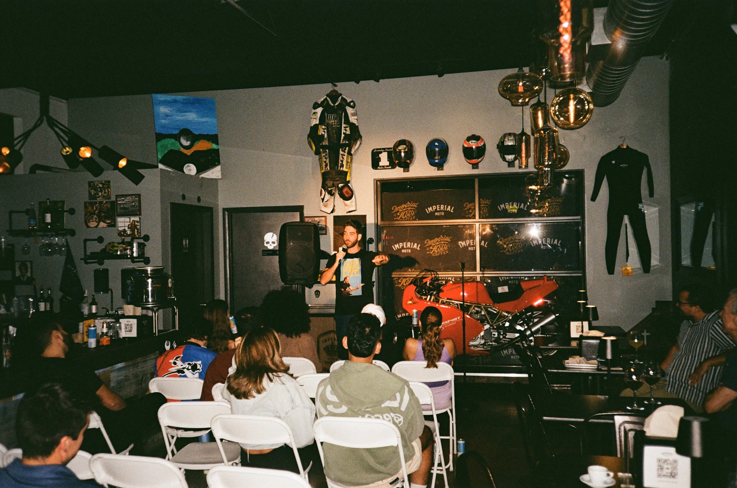 A man in a black T-shirt with a microphone stands on a stage in a dimly lit bar or cafe, addressing an audience seated in white folding chairs. The background features motorcycle helmets, a racing suit, and a red motorcycle. The walls are decorated w