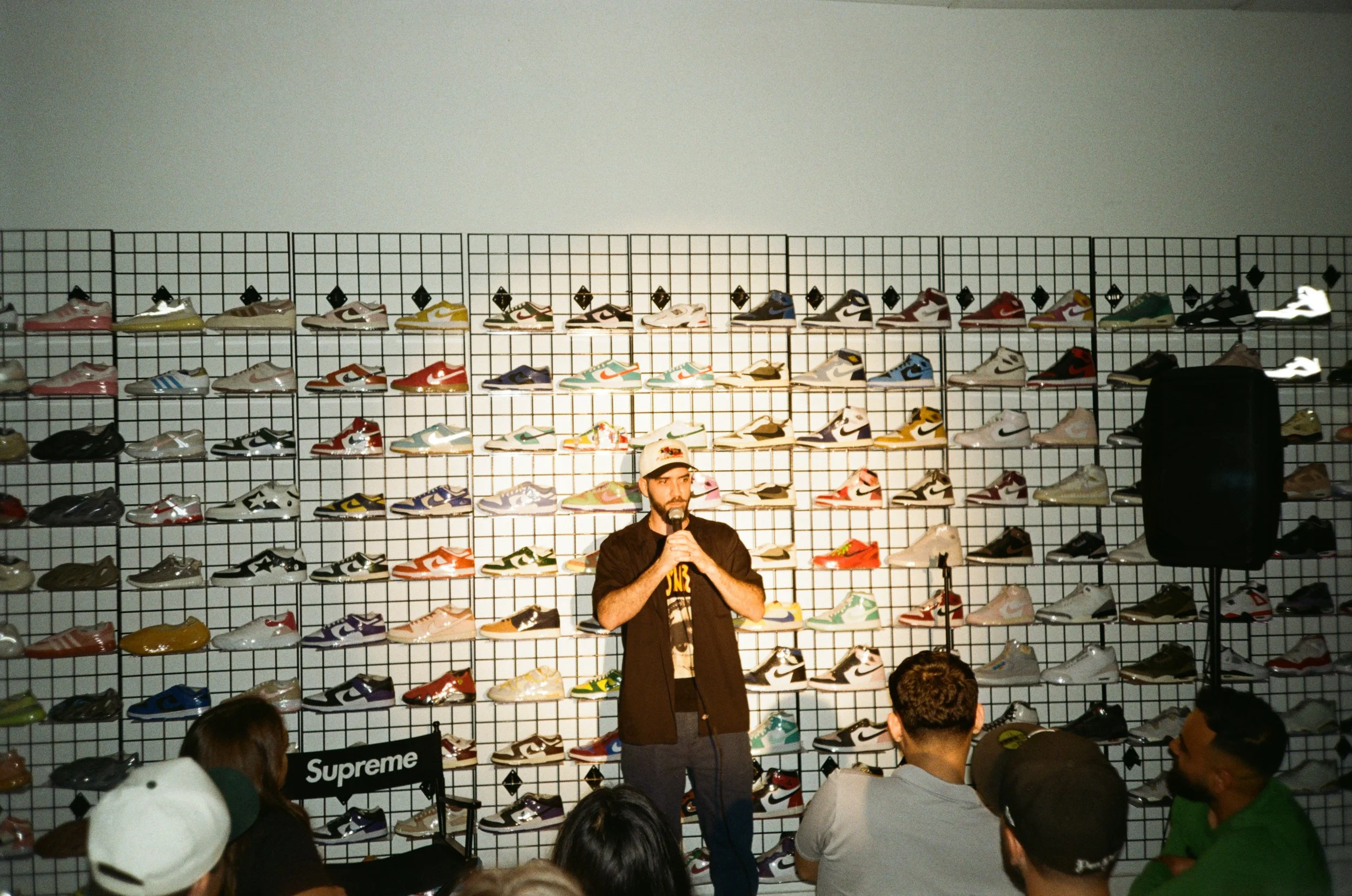 A man stands on a stage with a microphone in hand, speaking to an audience in front of a wall display of various sneakers. The display features multiple rows of sneakers, including brands like Nike and Adidas, arranged on a grid backdrop. There is a 