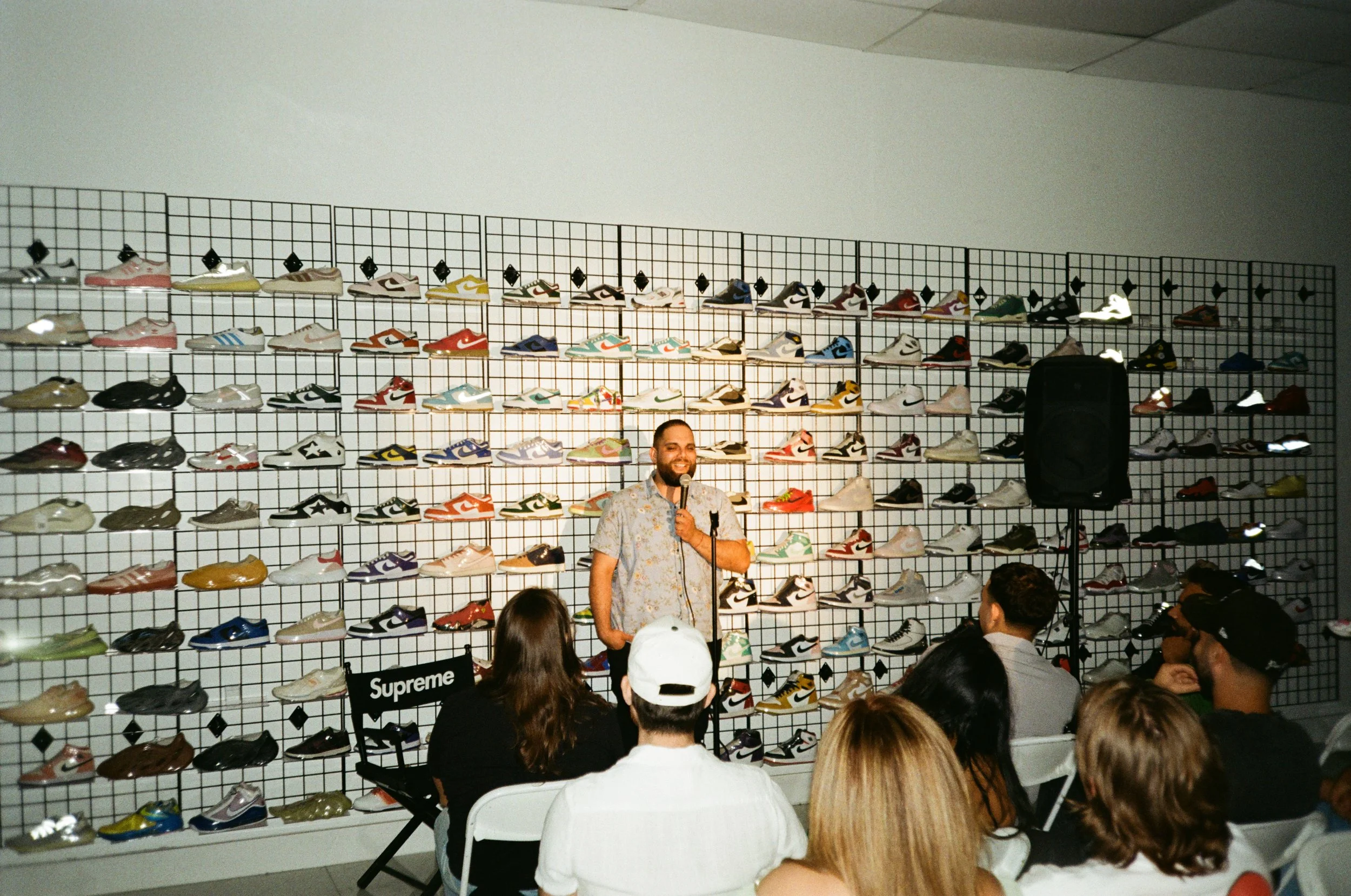 A man standing on stage with a microphone, speaking to an audience in front of a wall display of various sneakers. The sneakers are arranged on a grid wall, creating a colorful backdrop. The audience is seated, and some are looking at the speaker, wh