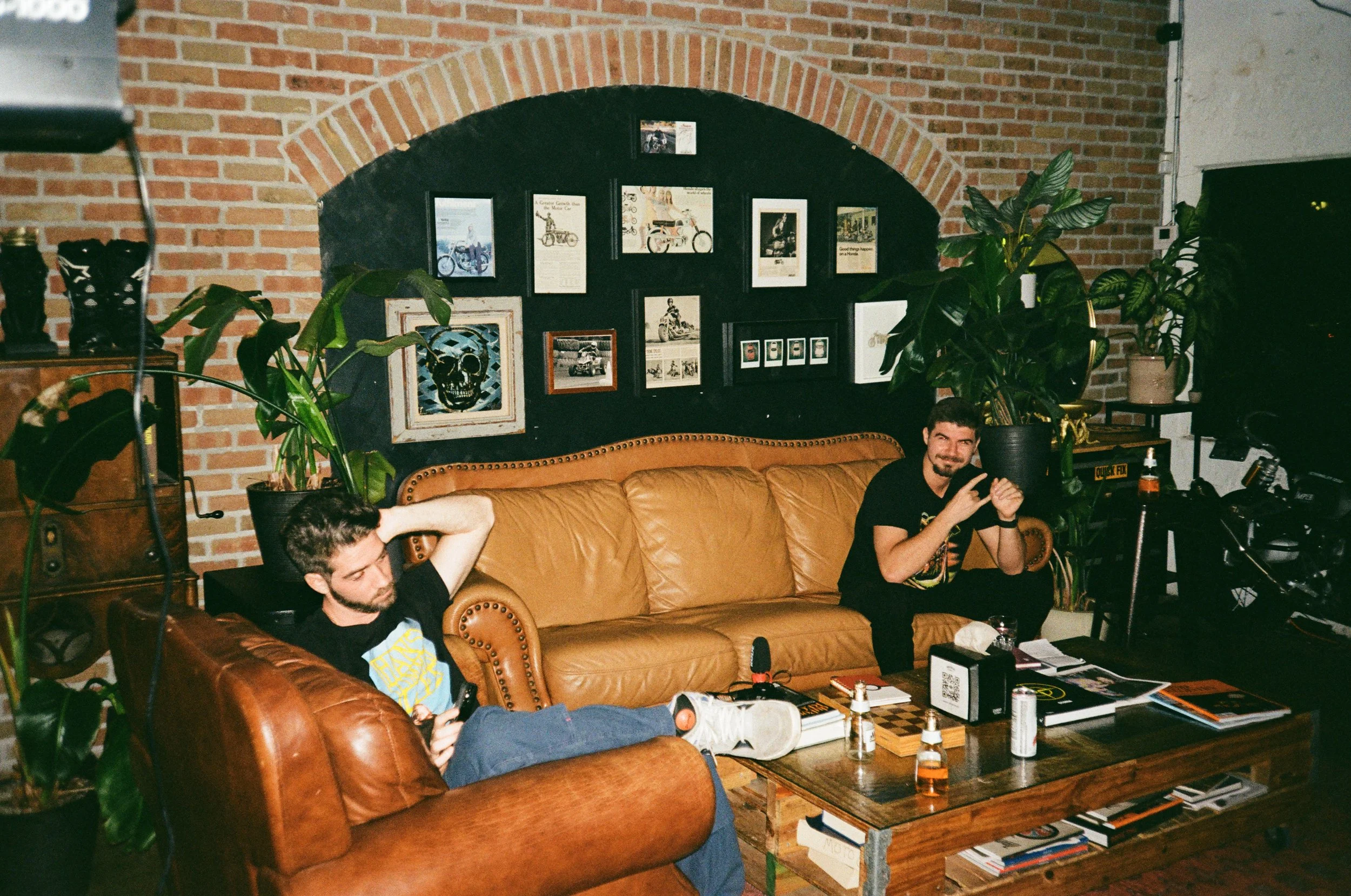 Two men sitting on leather sofas in a cozy, art-filled room with brick walls, plants, and various framed pictures and artwork on the wall.
