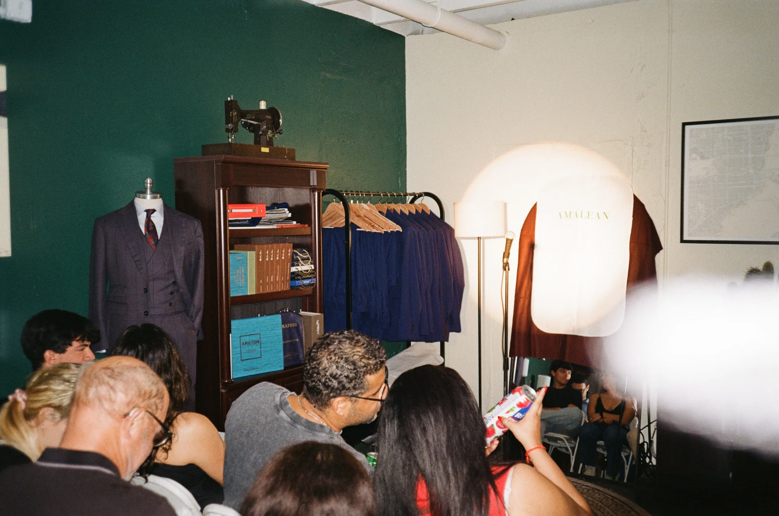 People attending a fashion show or presentation in a room with clothing racks and a mannequin dressed in a suit.
