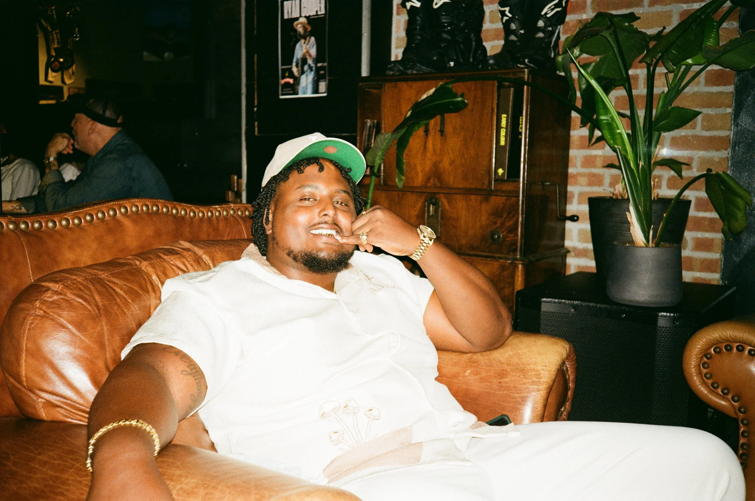 A man with a white cap, jewelry, and a white shirt is sitting on a tan leather couch in a cozy, artsy space, smiling at the camera. There are plants and vintage decor in the background.