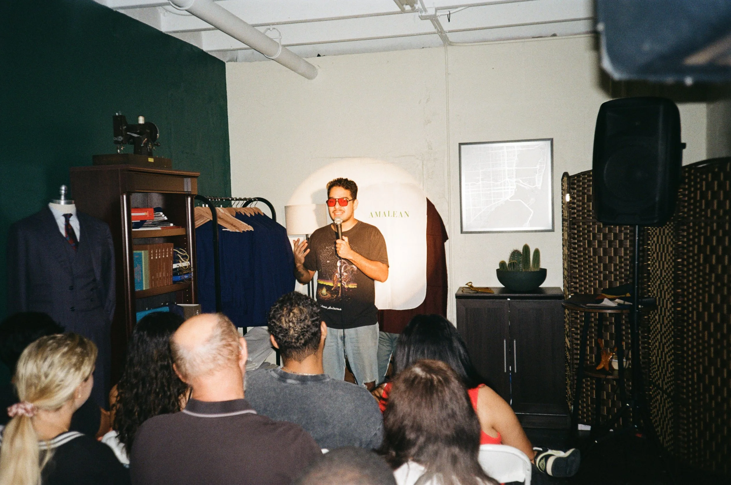 A man wearing red glasses and a graphic T-shirt is speaking into a microphone to an audience seated in front. The setting appears to be a small comedy club or event space with clothing racks, a suit on a mannequin, plants, and artwork on the wall.