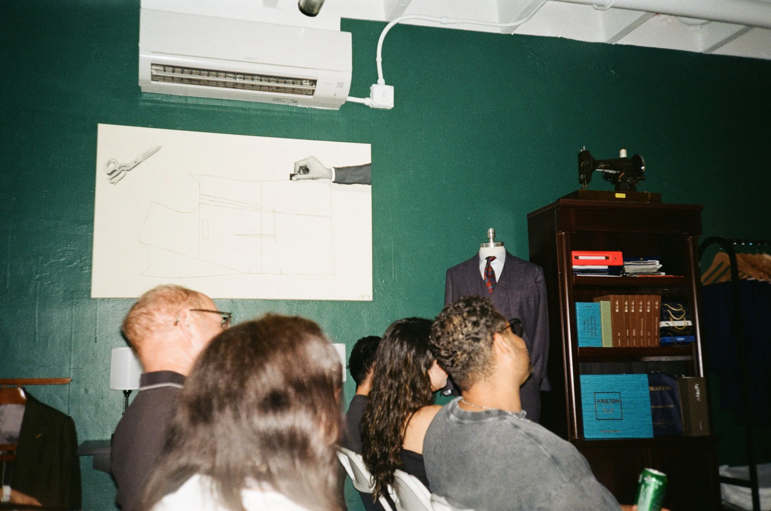 People attending a presentation or lecture in a room with a green wall, a whiteboard with sketches, a mannequin dressed in a suit, and a cabinet with books and an old sewing machine on top.