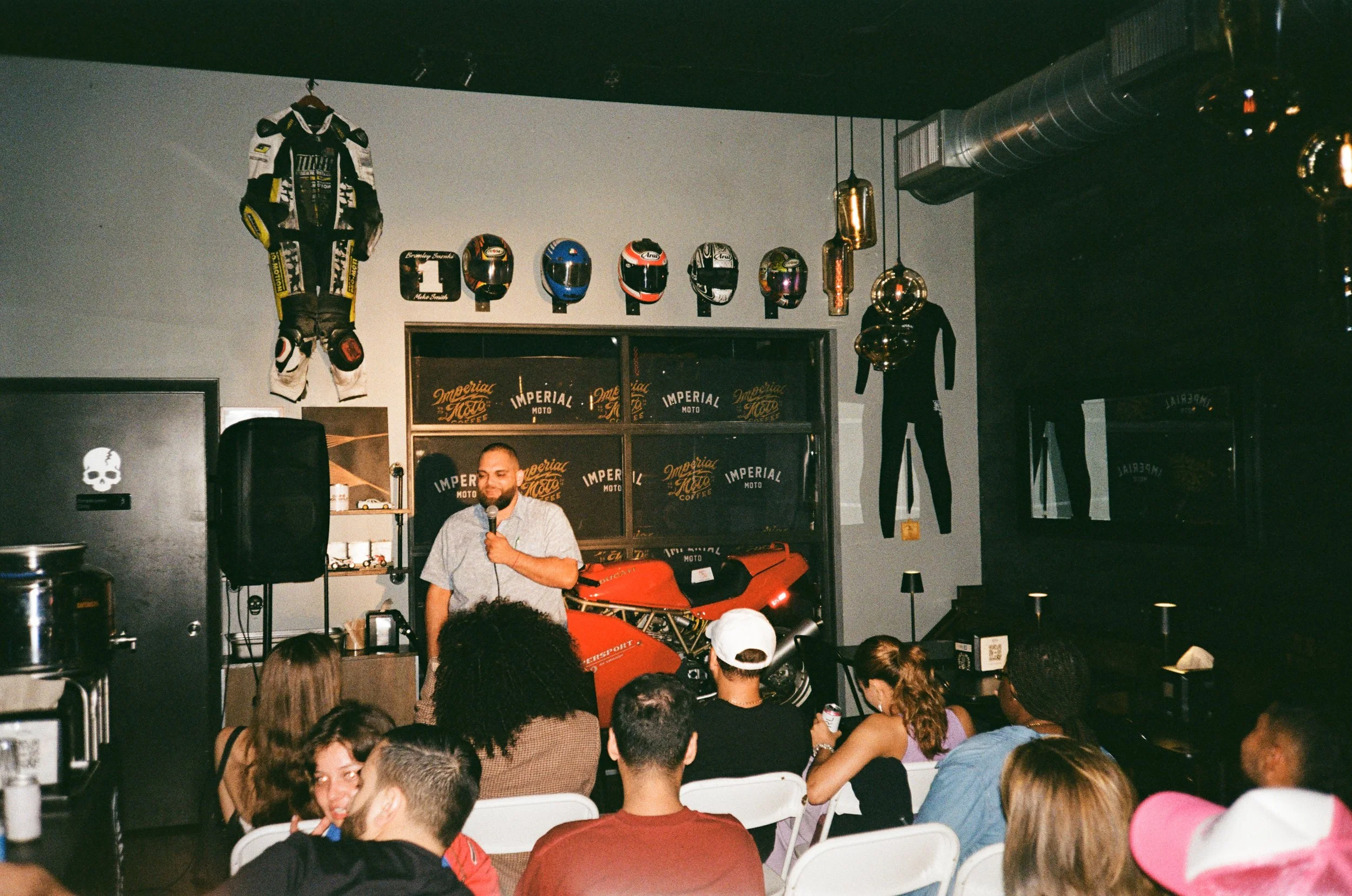 A man giving a speech or performance on stage in a room with motorcycle helmets and racing gear displayed on the wall behind him. There are audience members seated in front, and the room has a dark industrial style with hanging gold-colored pendant l