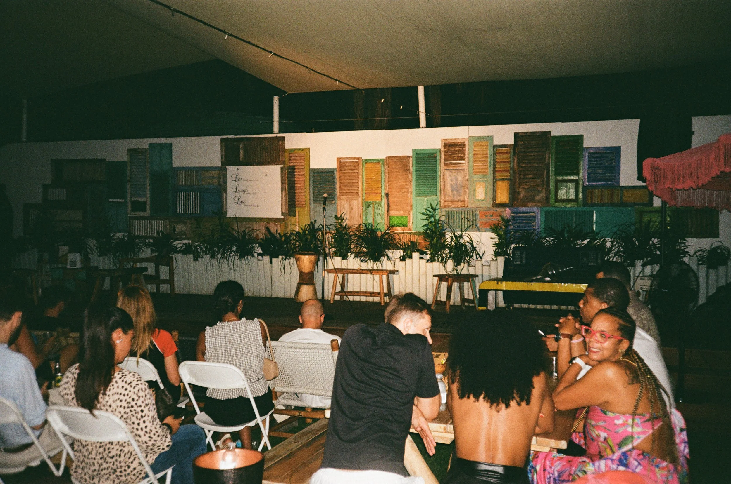 A group of people sitting and socializing at tables in a cozy, decorated indoor venue with a stage backdrop decorated with colorful wooden window shutters and plants.