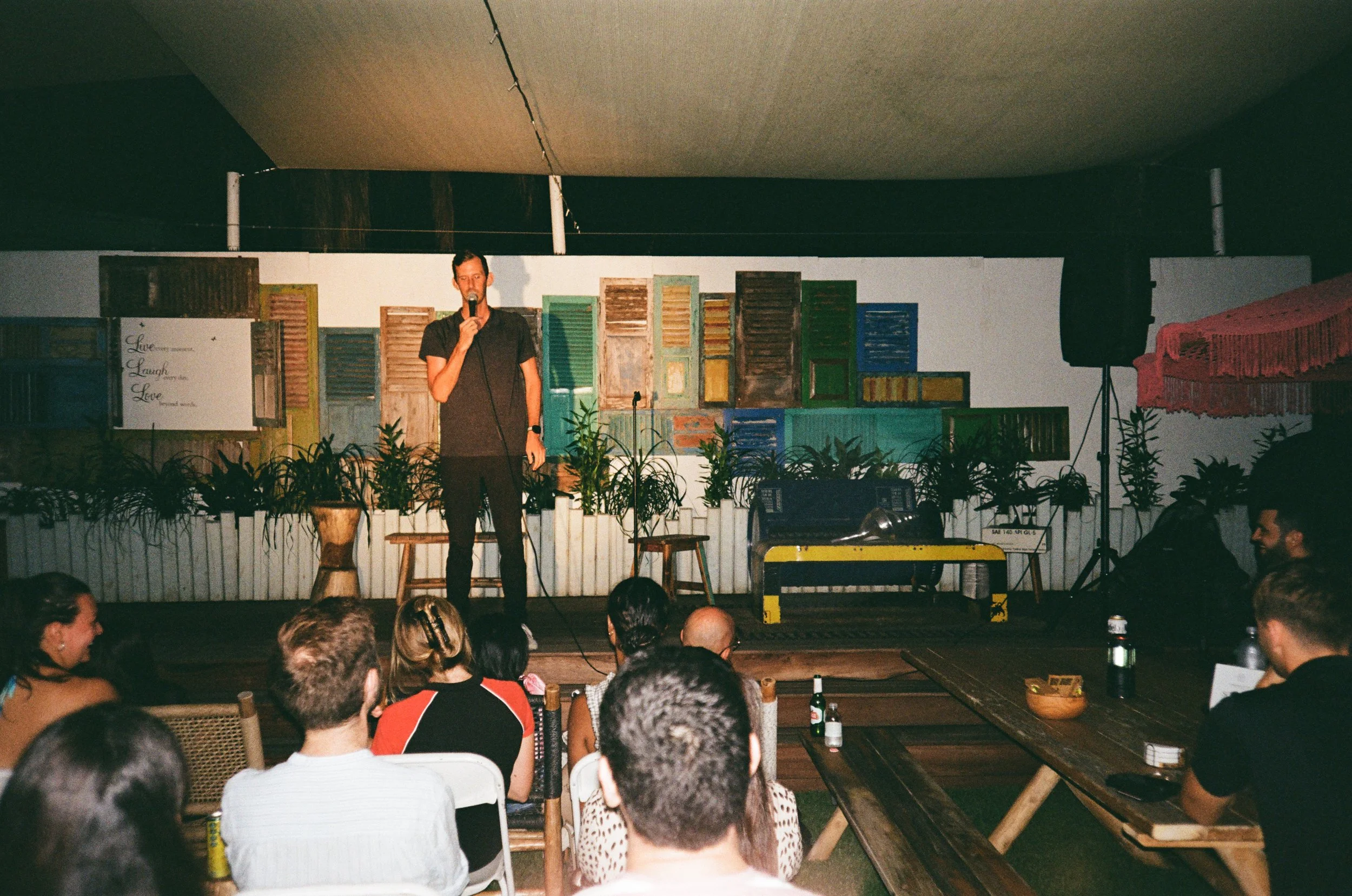 A man stands on a stage performing comedy in front of a seated audience. The stage has a backdrop of colorful, weathered shutters and plants, with a quote about love and laughter on the wall. Audience members are watching attentively, some with drink