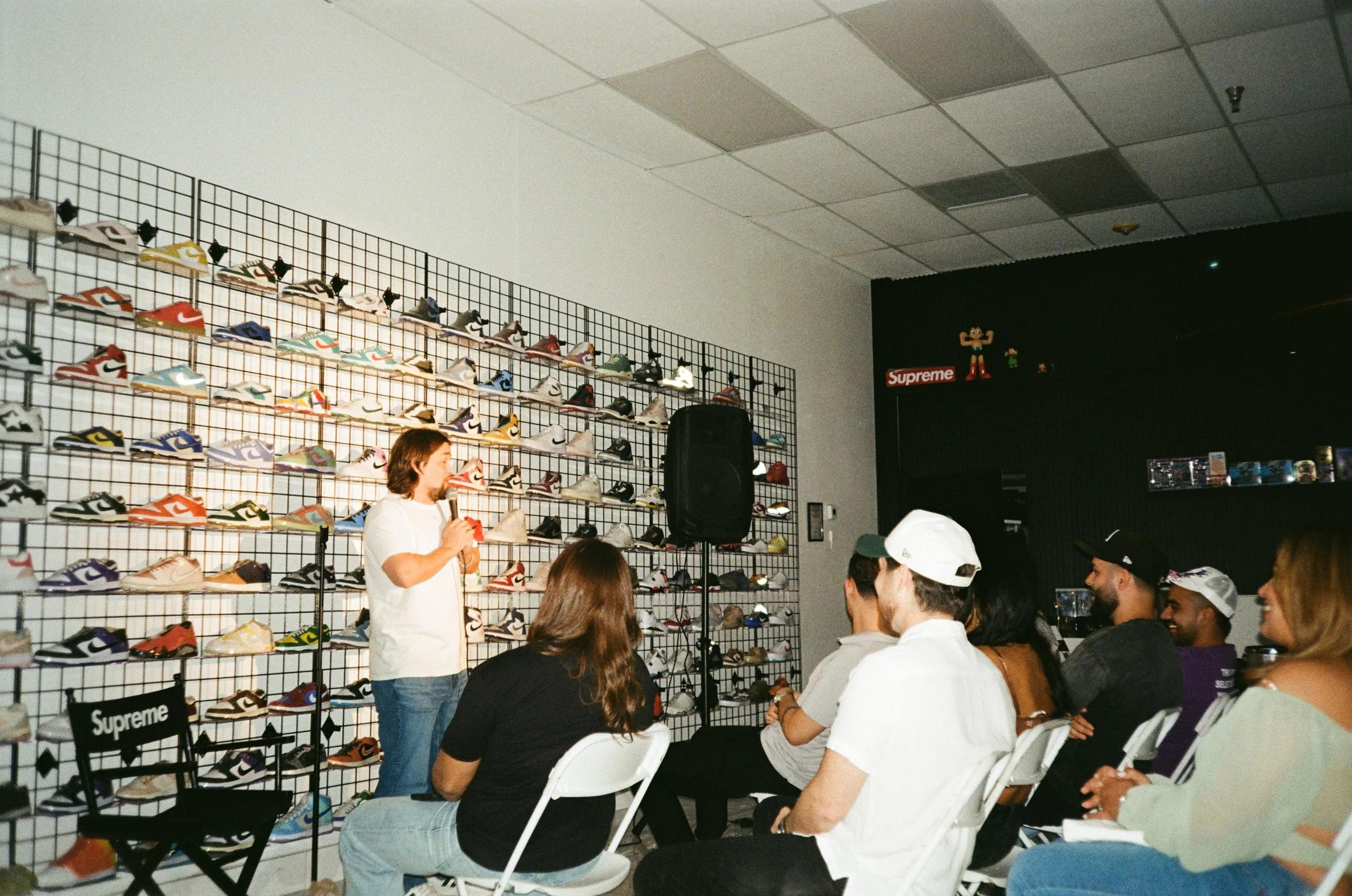 Group of people attending a presentation in a sneaker store. A woman is speaking into a microphone, standing in front of a wall with multiple pairs of sneakers on display. The audience is seated, watching the speaker, with some wearing caps.