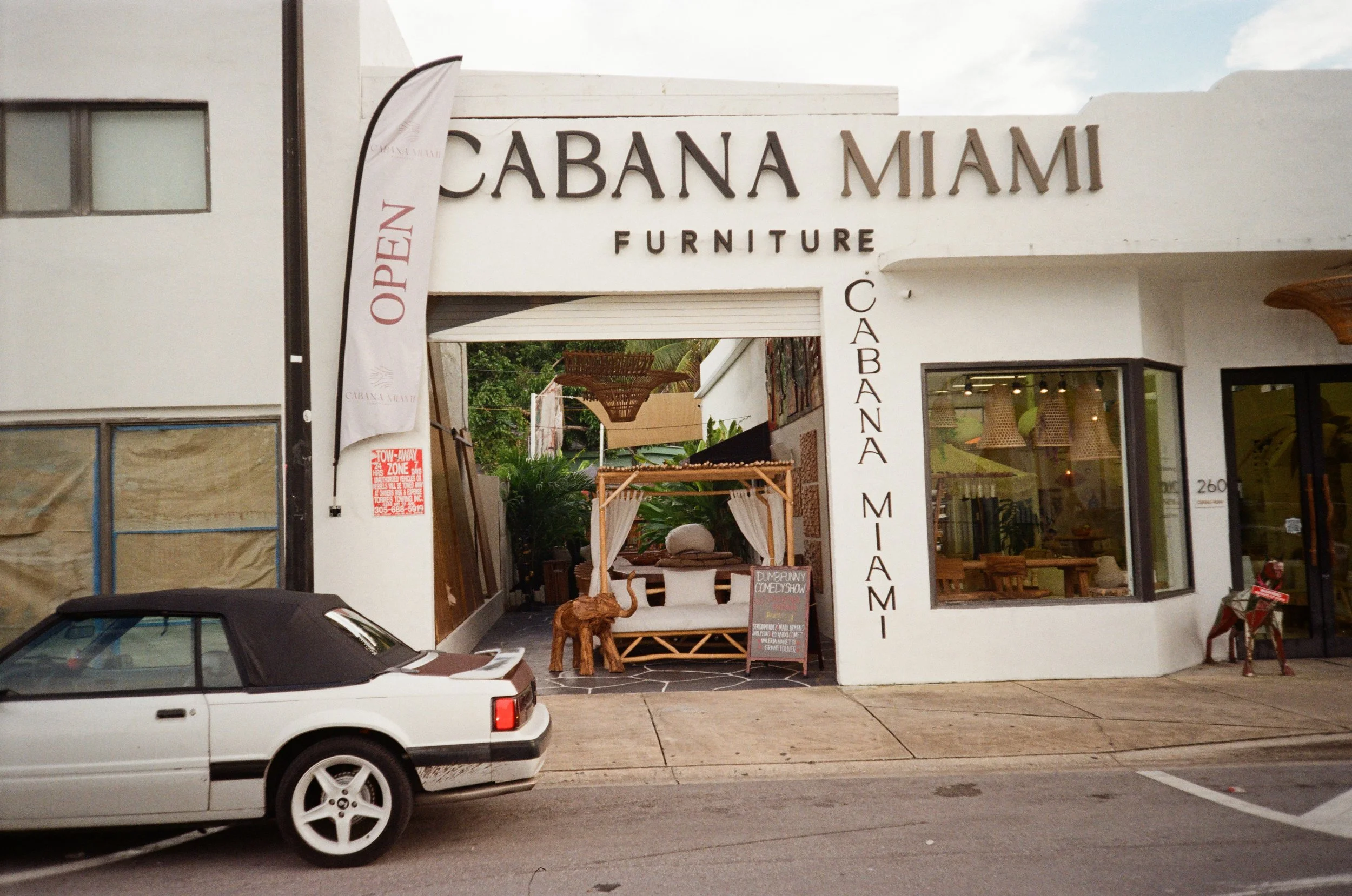 Exterior of Cabana Miami Furniture store with open sign, a white convertible car, and outdoor seating area with a canopy, elephant statue, and a signboard.