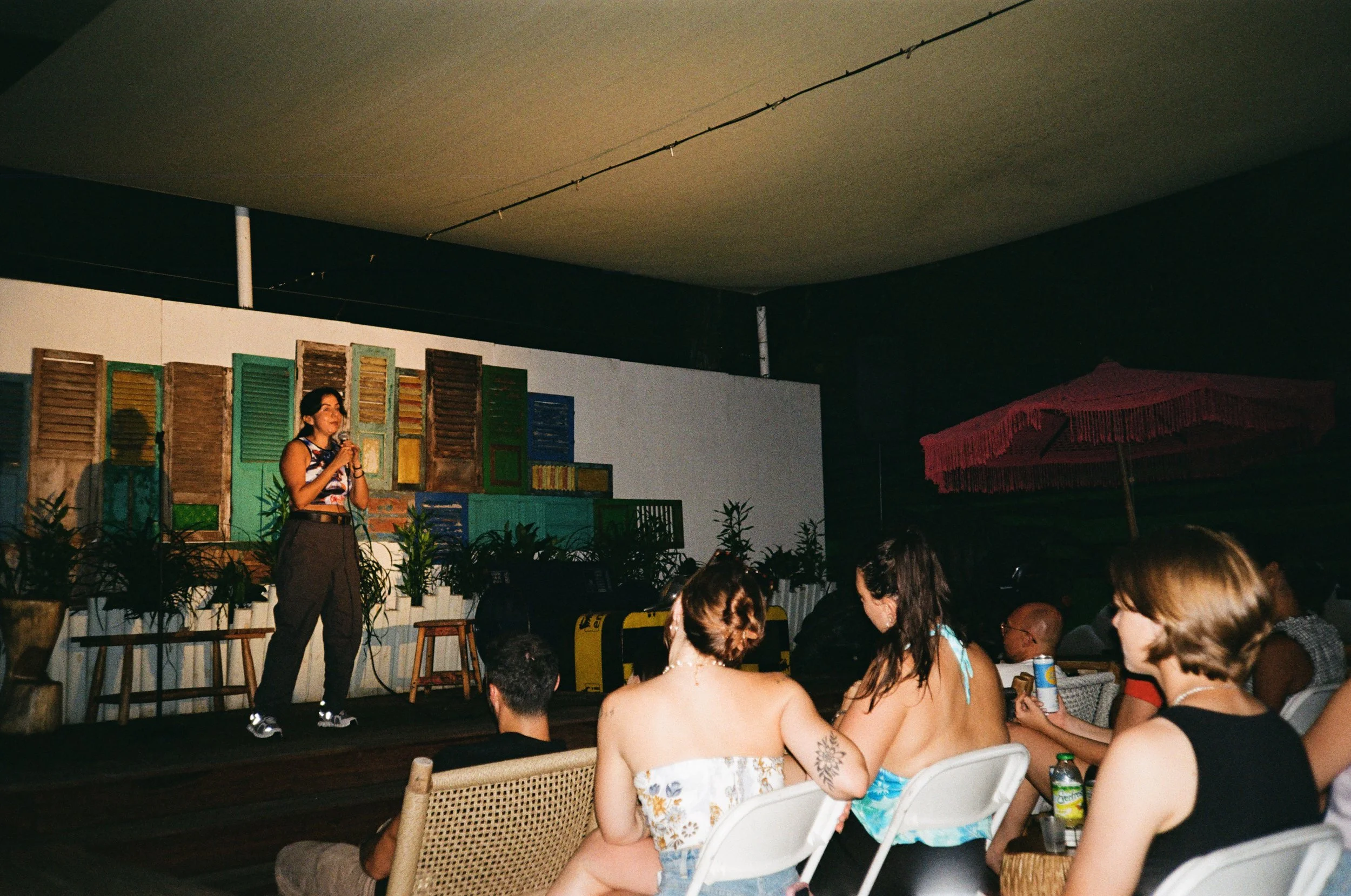 A woman performs on a stage with a colorful backdrop of wooden shutters, while a seated audience watches her, some with drinks, under a pink patio umbrella.