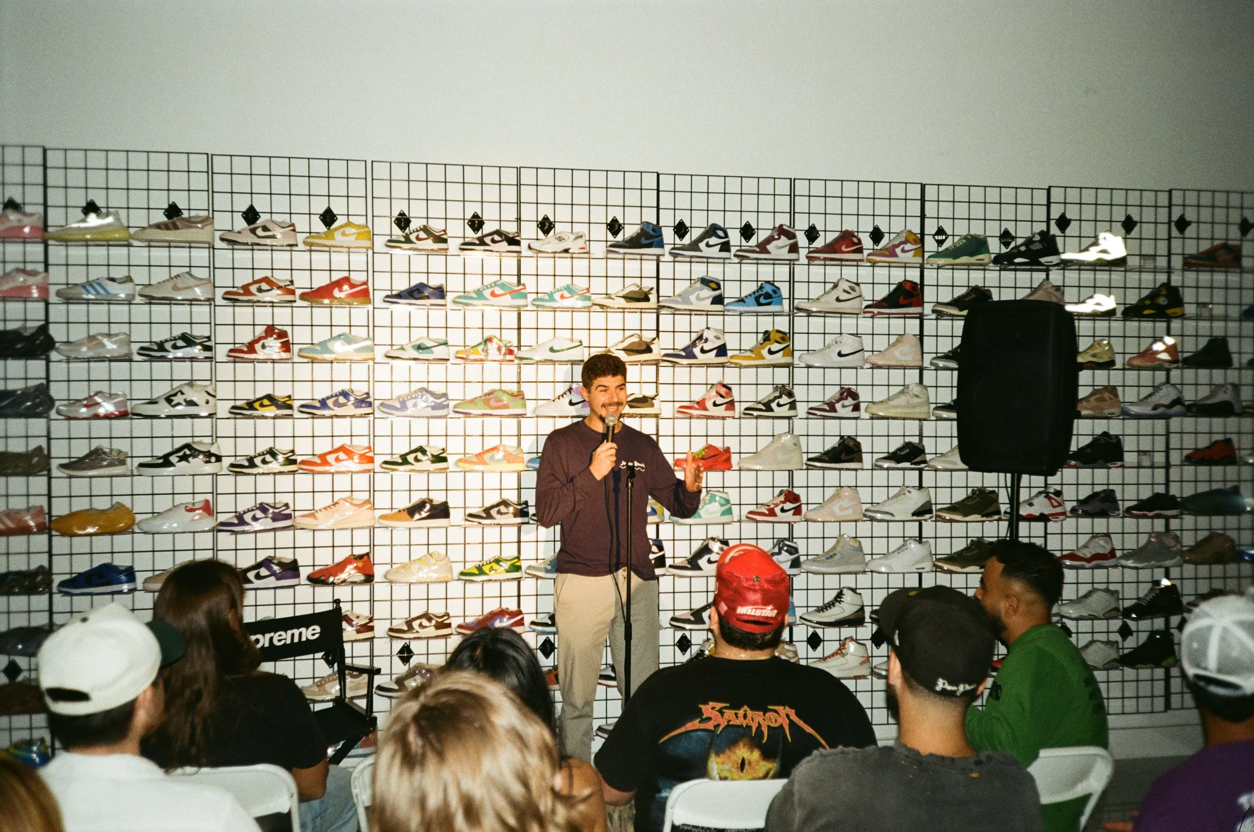A man standing in front of a wall display full of sneakers, giving a speech to an audience seated in front of him.