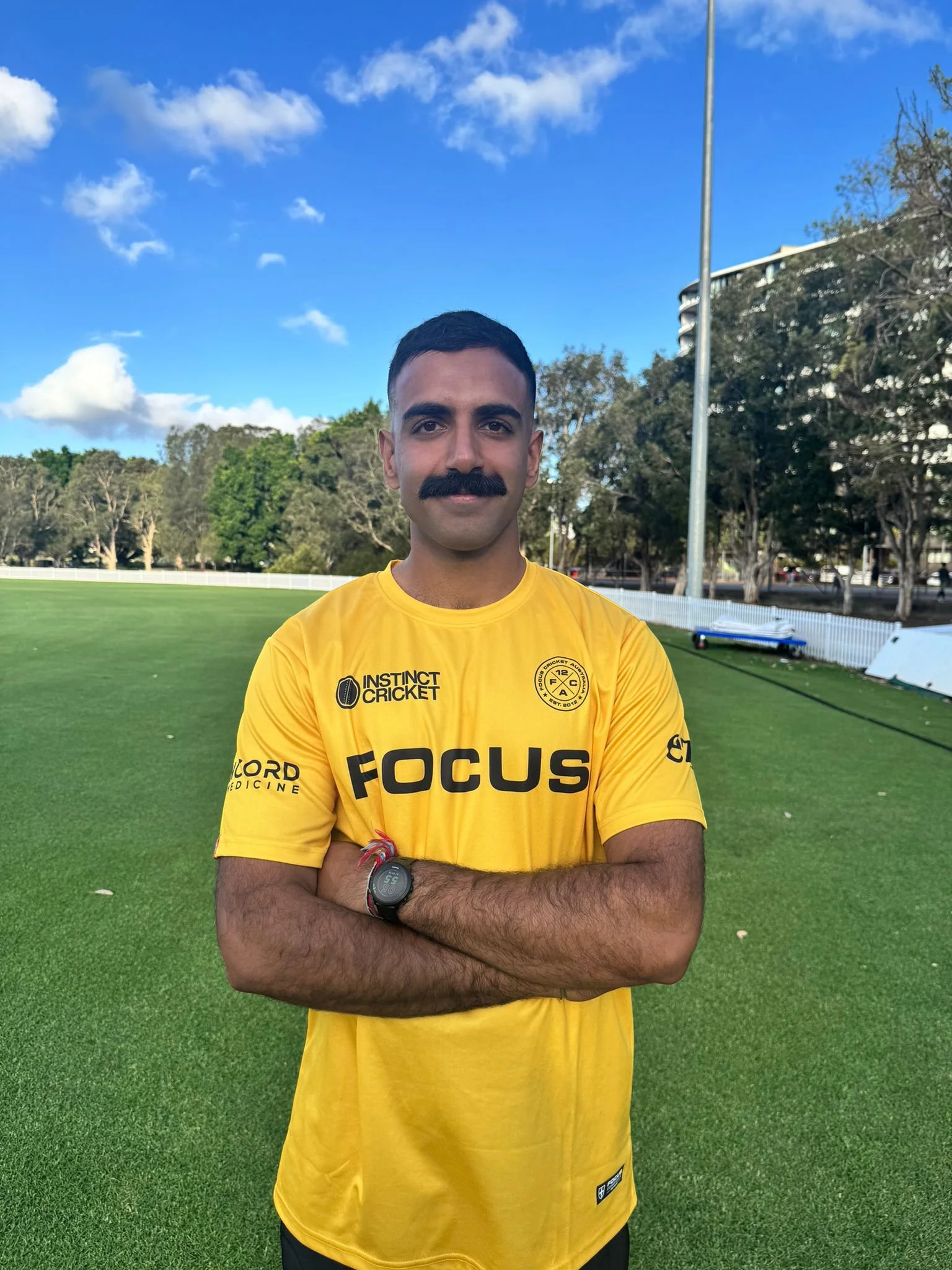 Man standing in front of a yellow flag with 'Instinct Cricket' logo and text, wearing a cricket t-shirt and cap, arms crossed, outdoors on a sunny day.