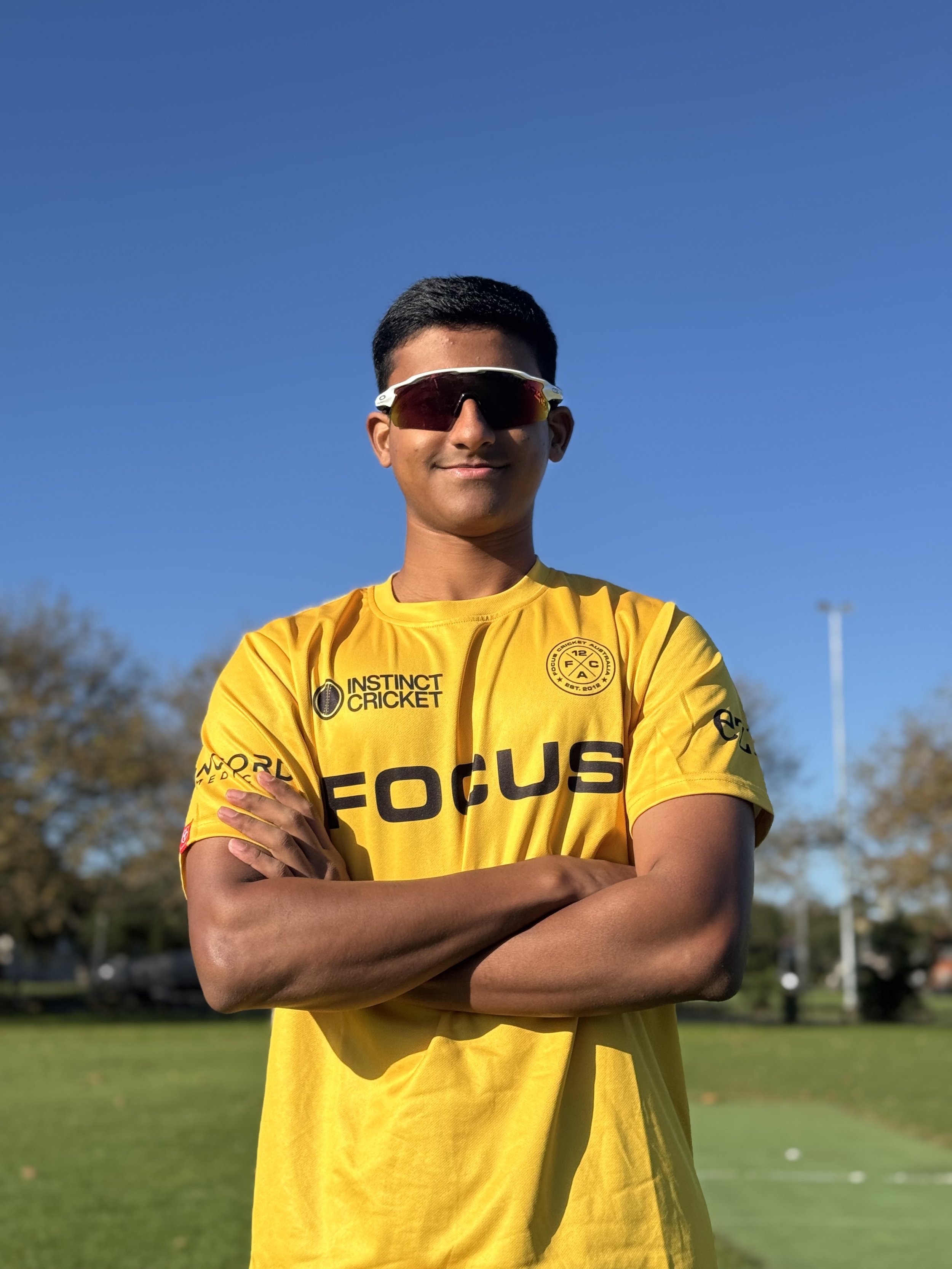 A young man with curly brown hair wearing a yellow sports jersey with logos, standing outdoors with trees and bushes in the background.