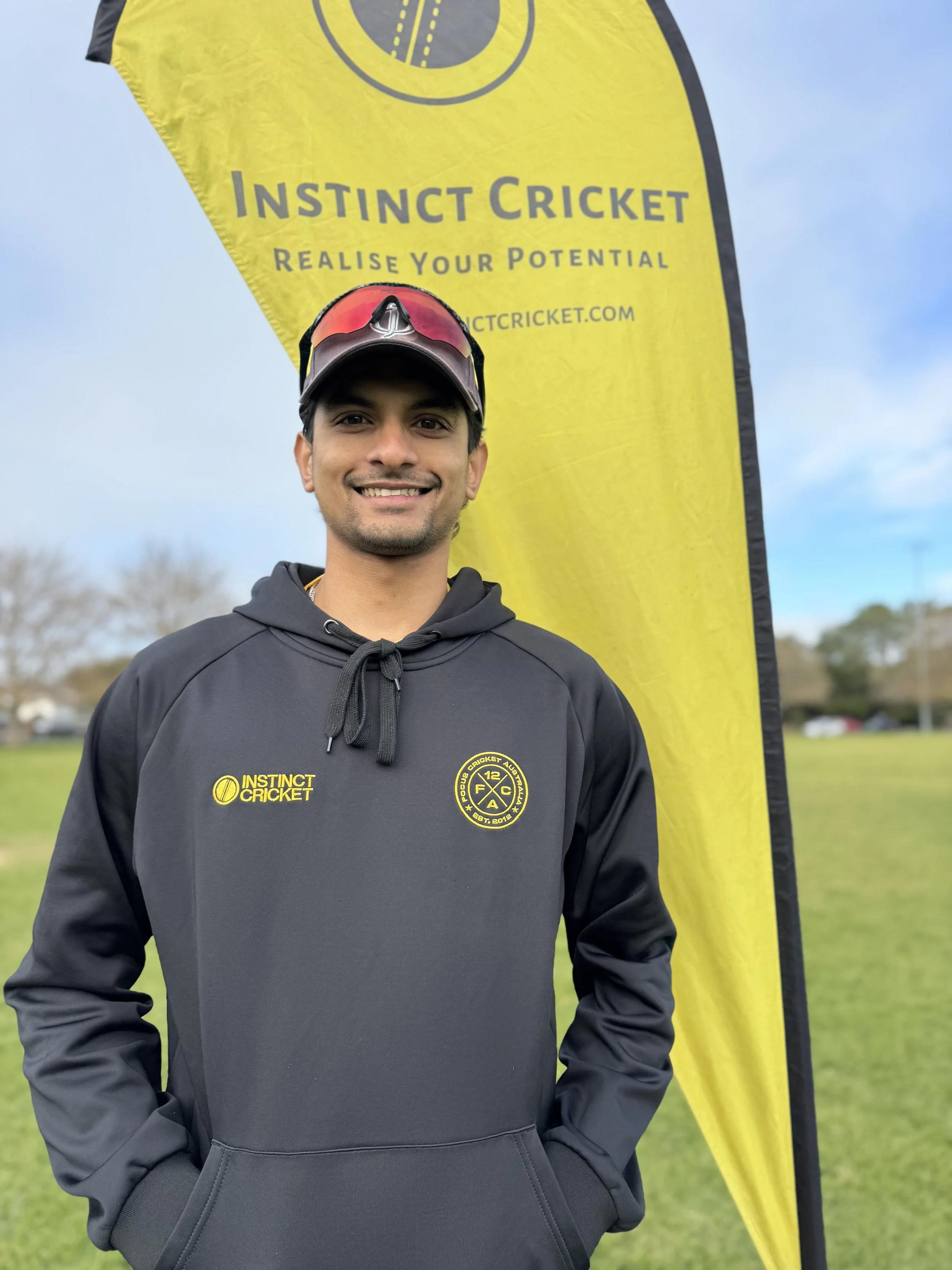 A young male athlete in a white sports shirt and cap standing outdoors with arms crossed, in front of a yellow flag that reads 'Instinct Cricket' and 'Realize Your Potential.'