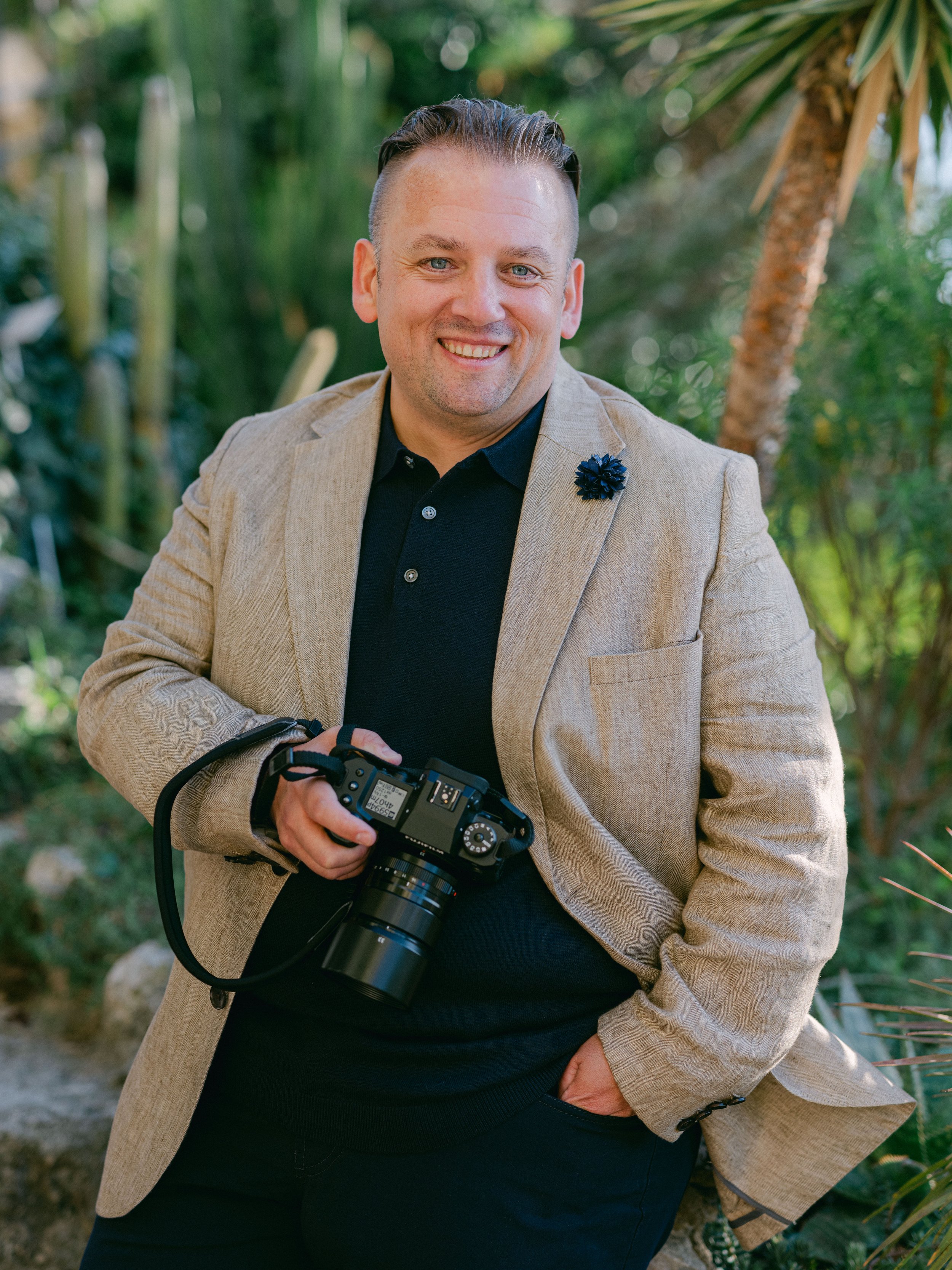 A man smiling and holding a camera in an outdoor setting with greenery and trees.