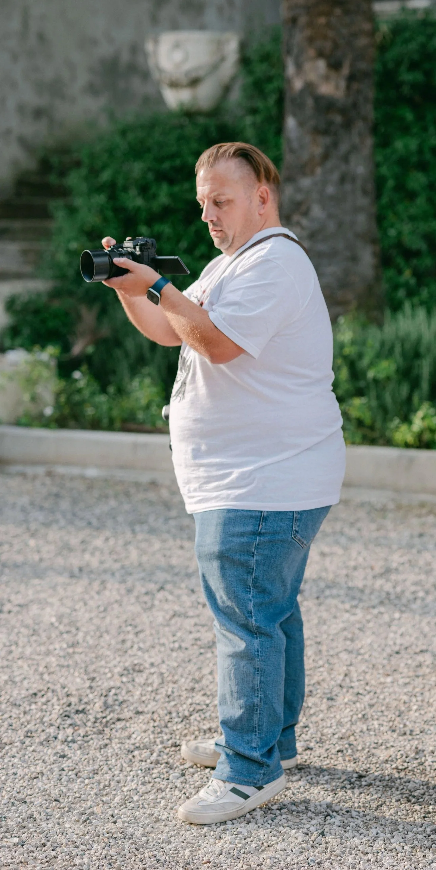 A man standing on gravel ground, wearing a white t-shirt, jeans, and white sneakers, is looking at a camera he is holding with both hands, outdoors with trees and greenery in the background.