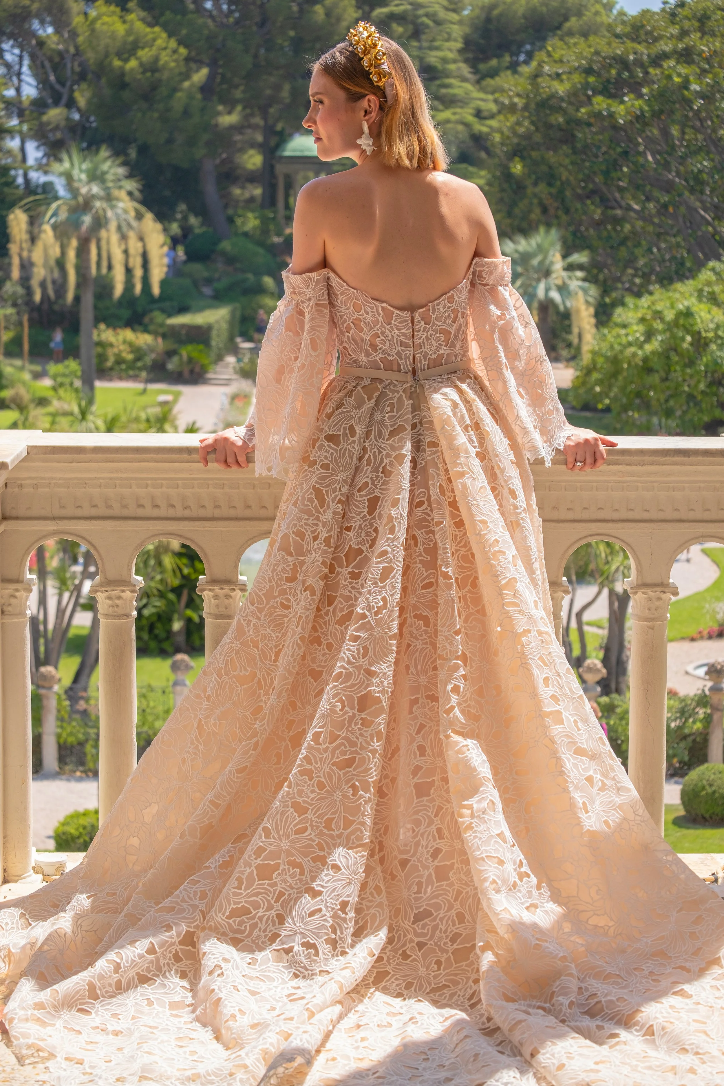 A woman wearing a light pink off-shoulder lace wedding dress with a train, standing on a balcony overlooking a lush, green garden at Villa Ephrussi.