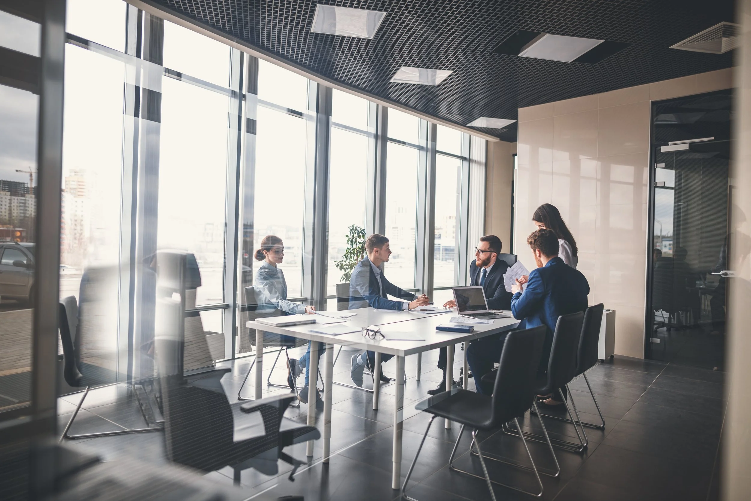 Business meeting in a modern office with large windows, five professionals sitting around a white table, two women and three men, engaged in discussion, with laptops, documents, and glasses on the table.