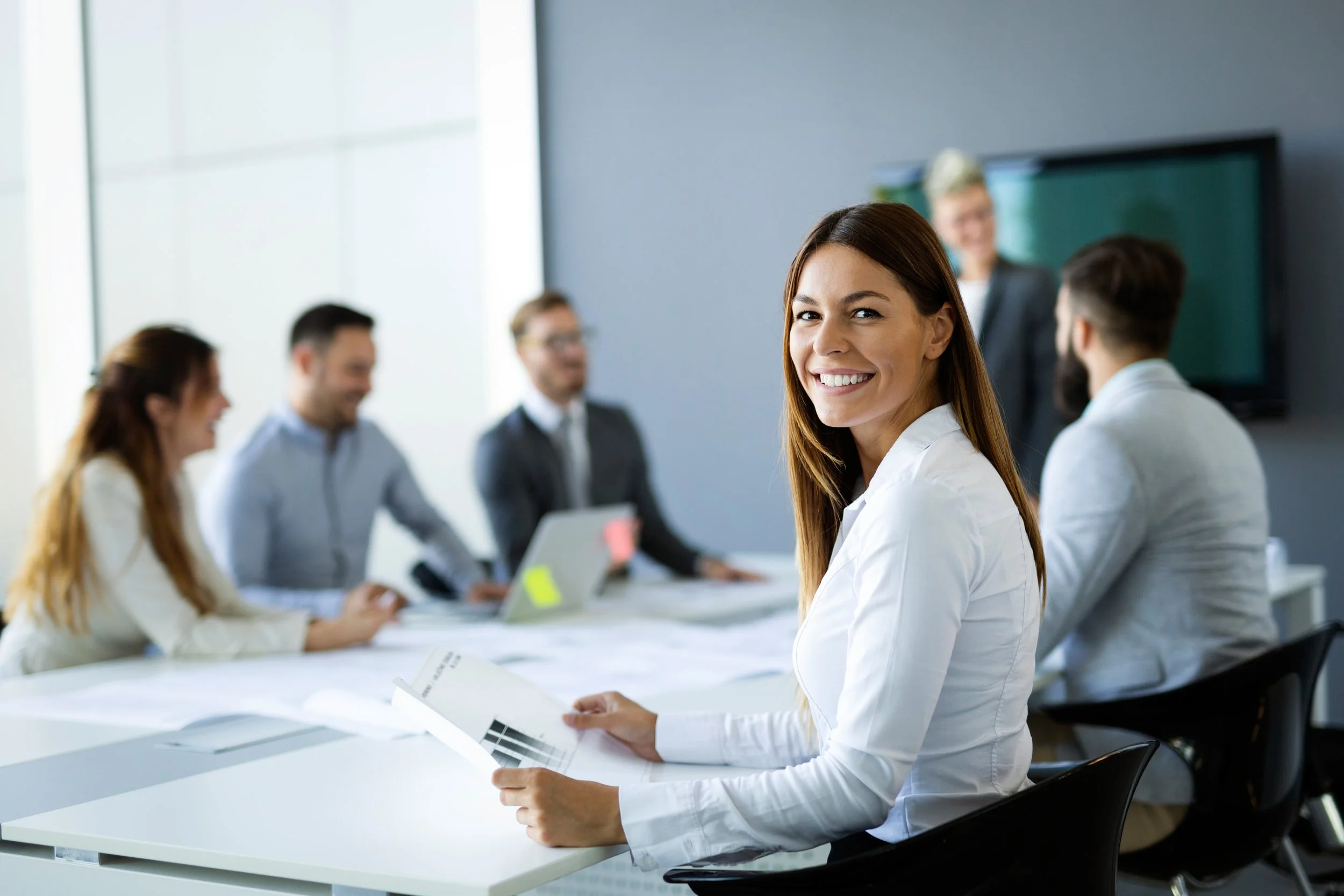 A group of business people in a meeting room, with a woman smiling and sitting at the forefront holding papers, and others engaged in conversation in the background.