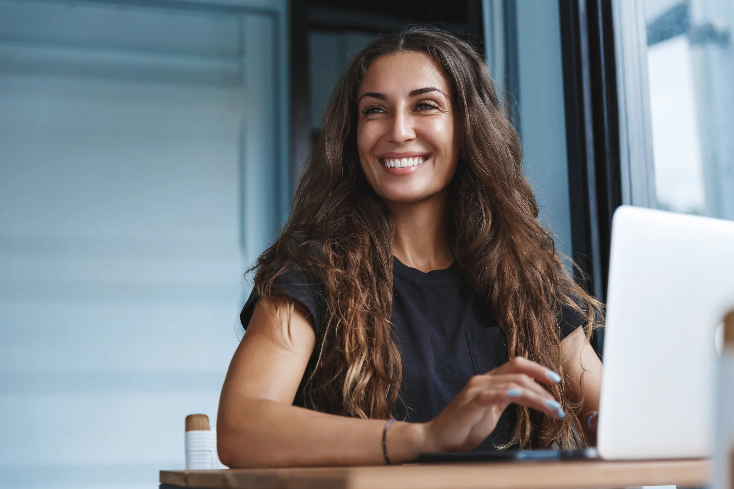 A young woman with long, wavy brown hair smiling while using a laptop at a desk near a window.