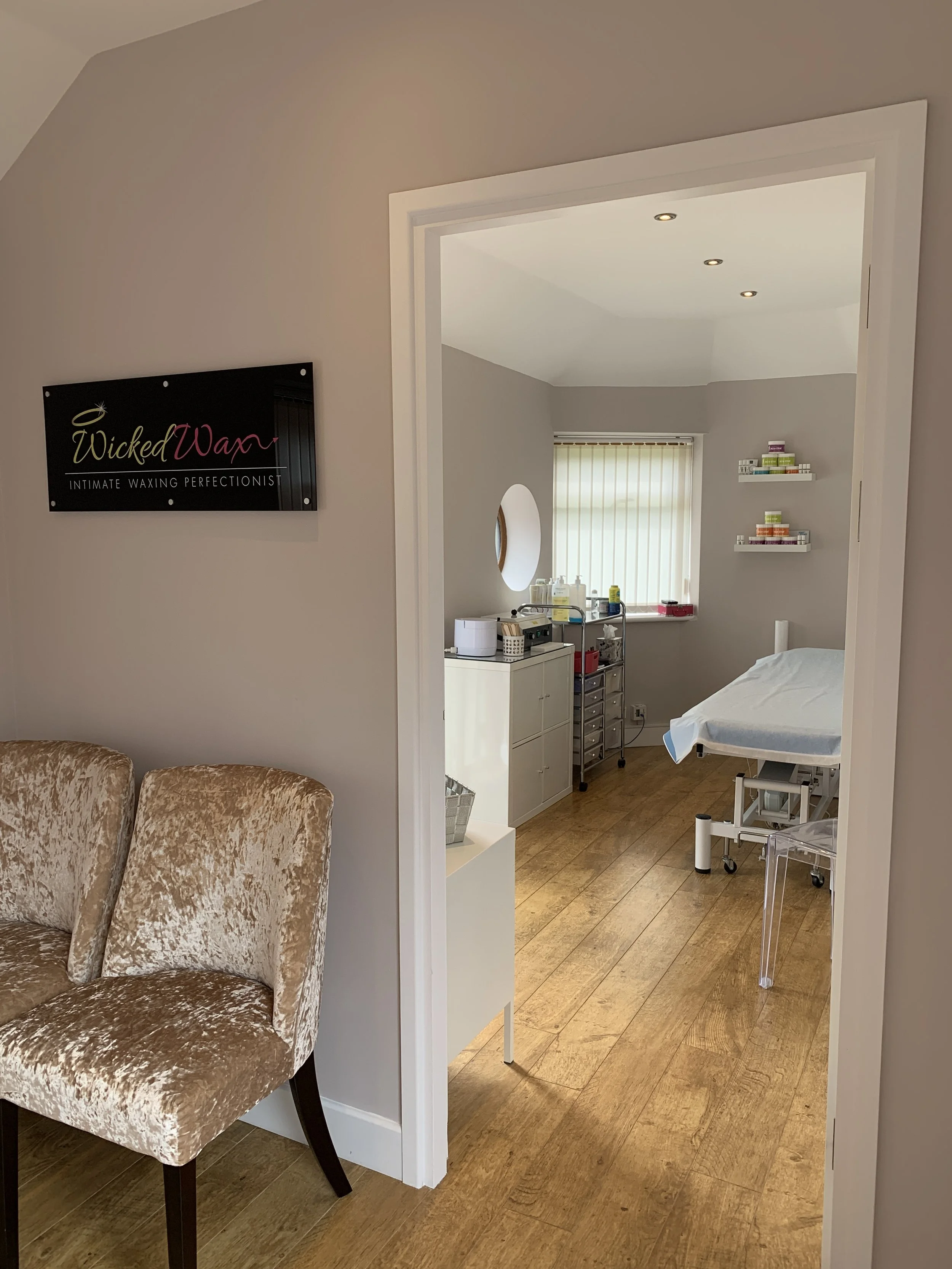 The image shows a room with a beige velvet armchair at the left, a sign on the wall that reads 'Wicked Wax - Intimate Waxing Perfectionist,' and a doorway leading to a treatment room with a white bed, shelves with colorful products, a cart with supplies, and a window with vertical blinds.