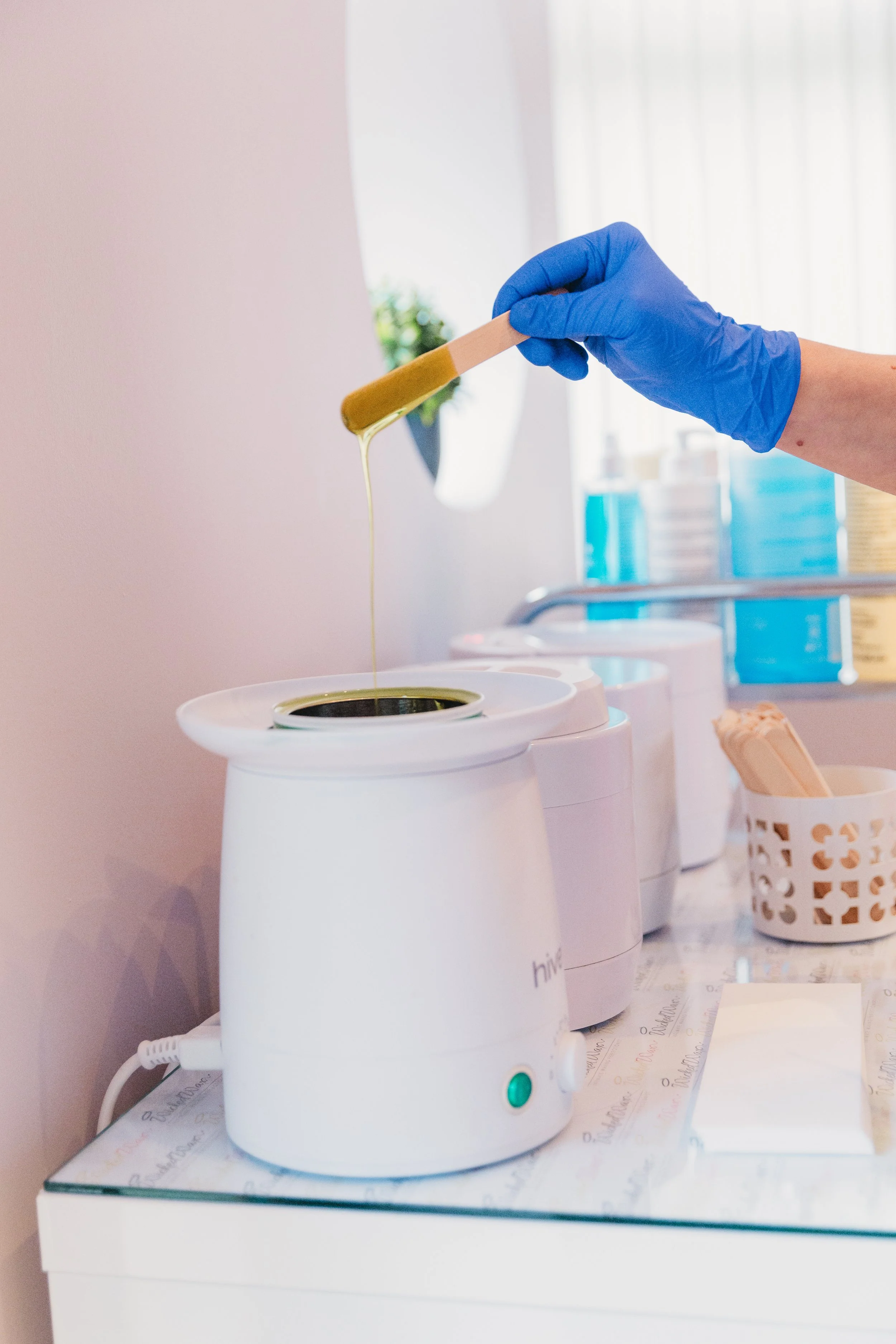 A person with a blue glove holding a wooden stir stick with green wax or resin dripping into a small white machine, possibly a wax warmer or melter, on a white counter.