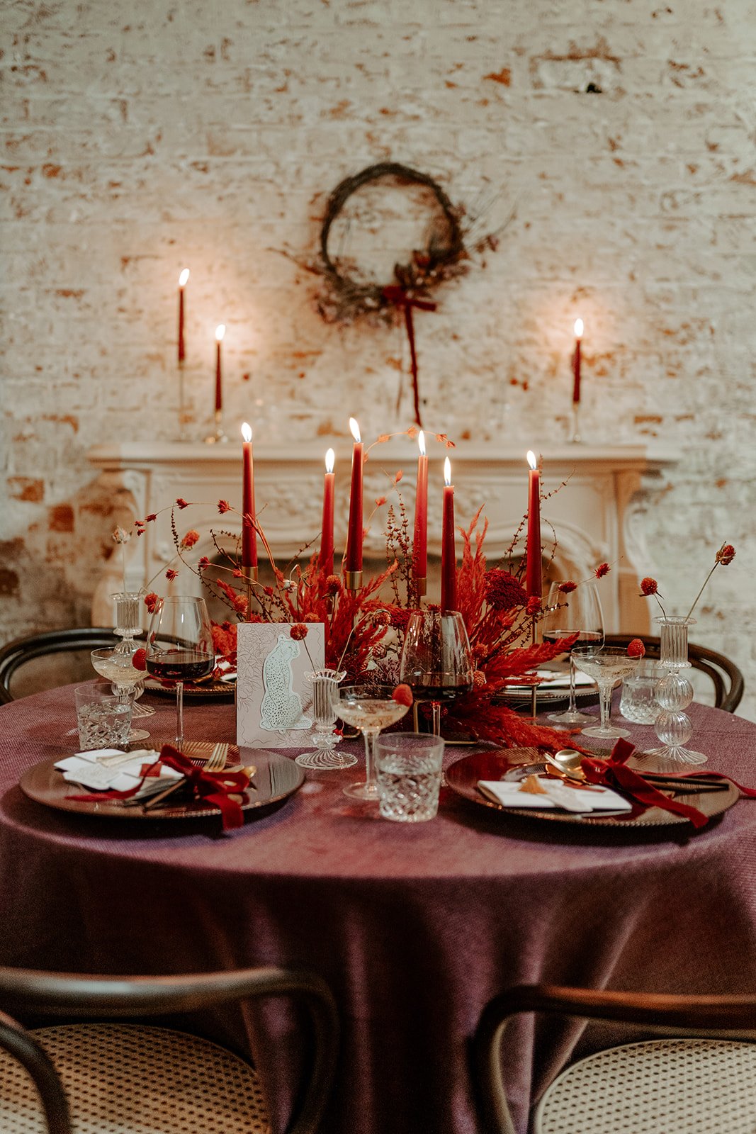 Elegant dining table decorated with red candles, floral centerpieces, and glassware, set against a brick wall with a wreath and additional candles on a mantel.