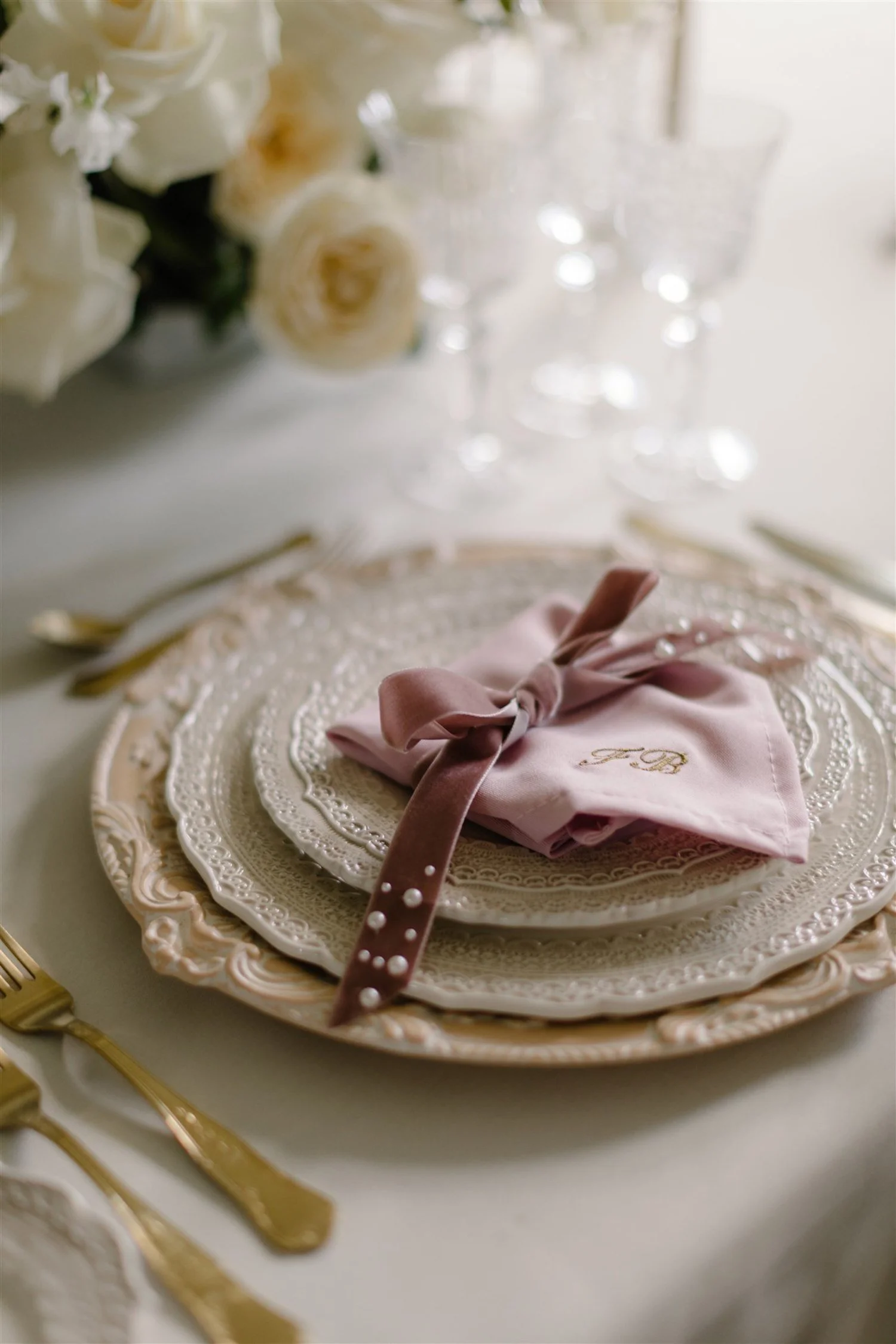 Elegant table setting with stacked ornate plates, a pink napkin tied with a ribbon, crystal glasses, and gold silverware; decorated with white roses.