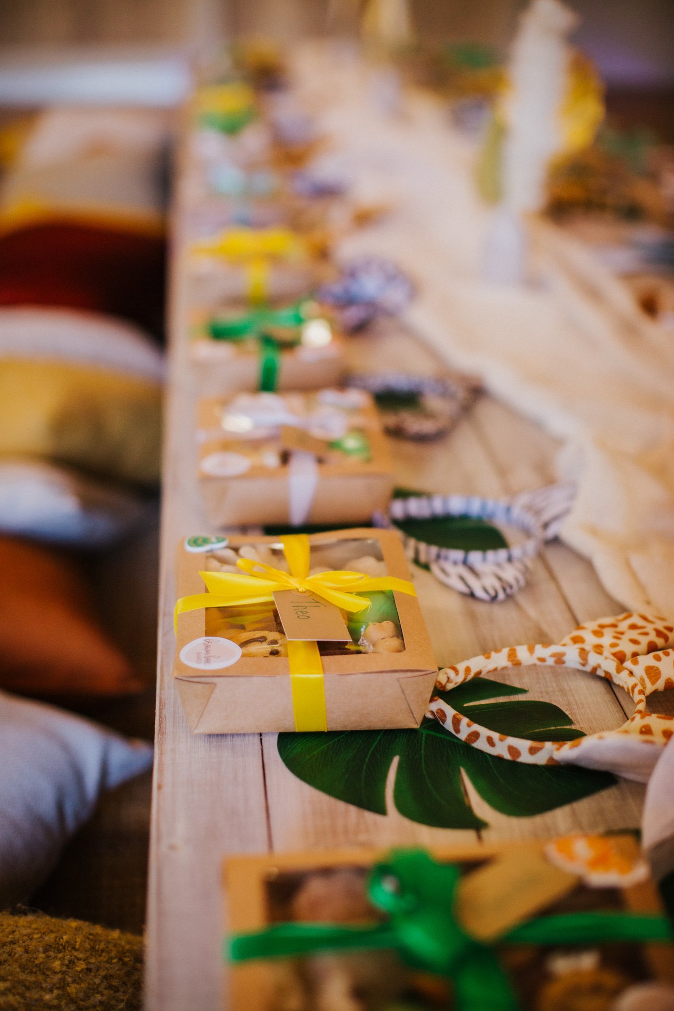 Table with Safari picnic boxes wrapped in yellow and green ribbons, surrounded by patterned cloths and green leaves.