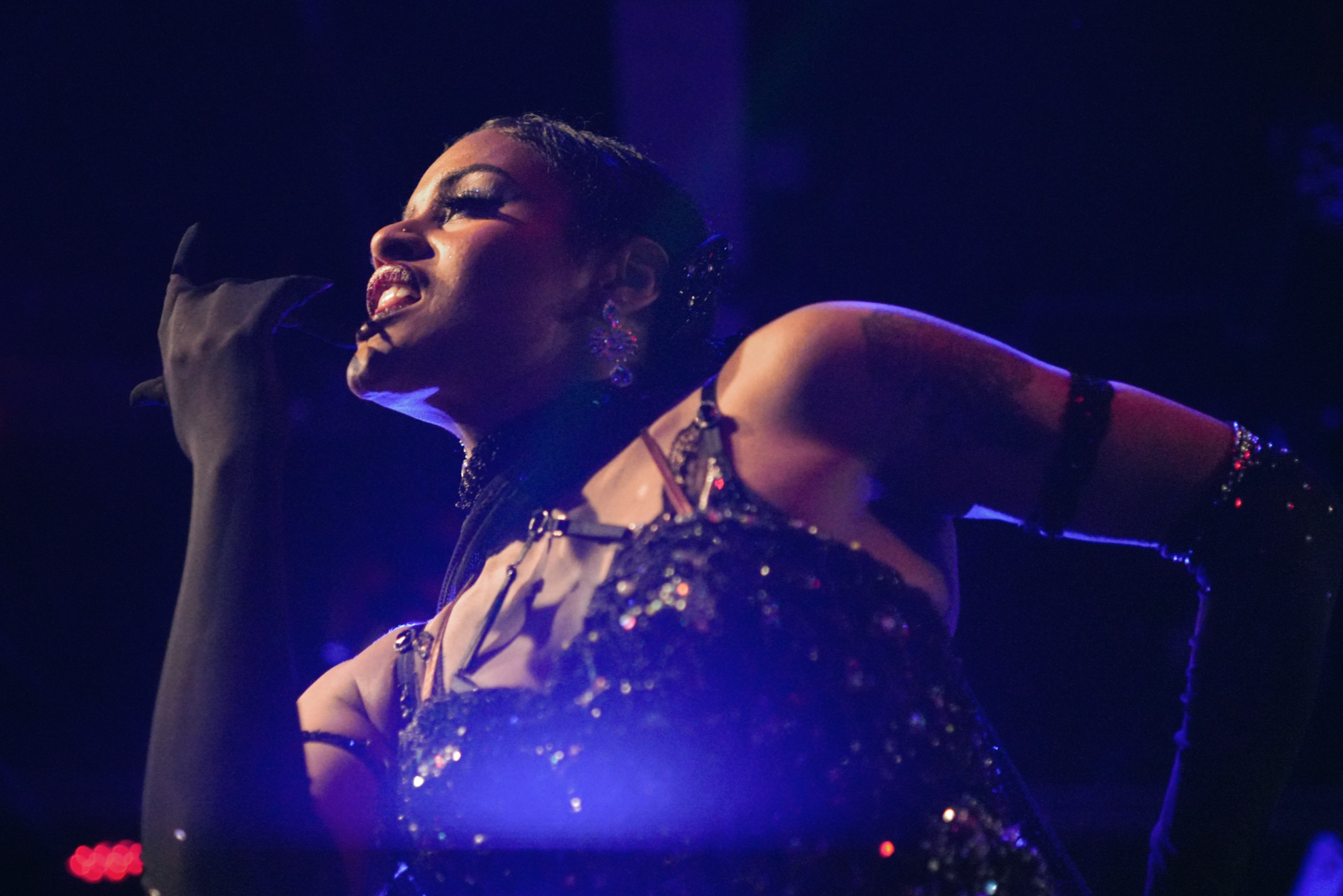 A woman singing into a microphone at an indoor event, wearing a silver sequined top, pink earrings, and a hair accessory, with people blurred in the background.