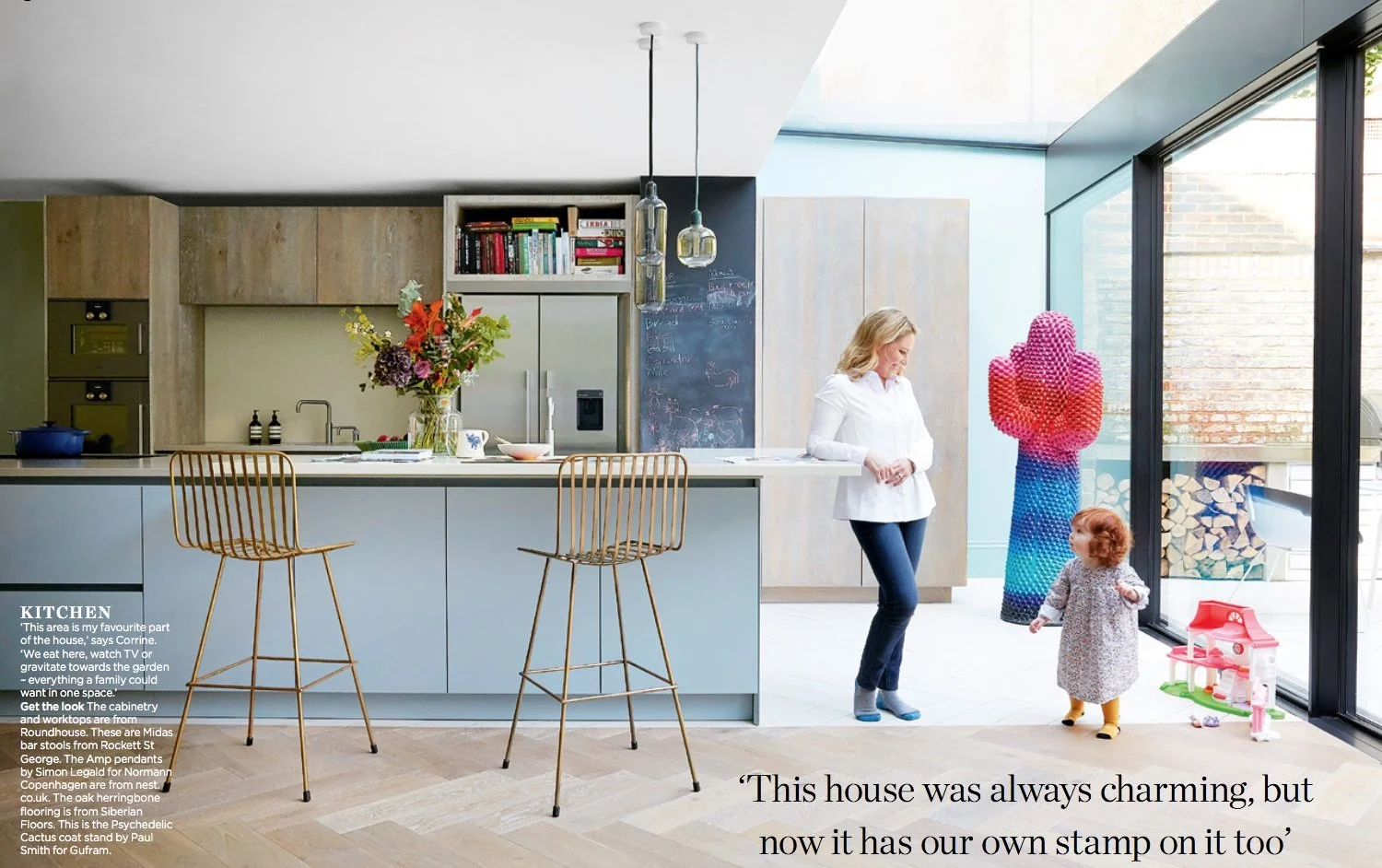 Modern kitchen interior with island, bar stools, and pendant lights; woman leaning on counter, toddler standing by a colorful cactus sculpture; large windows revealing outdoor view.
