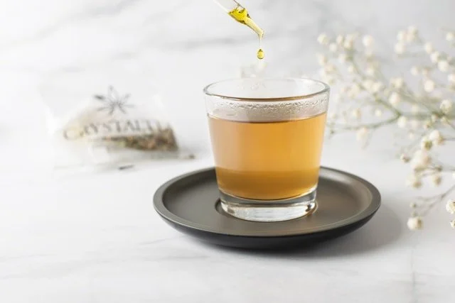 A glass of golden-hued tea or tincture on a saucer, featuring a dropper adding a single drop of oil, suggesting a calm and therapeutic setting.
