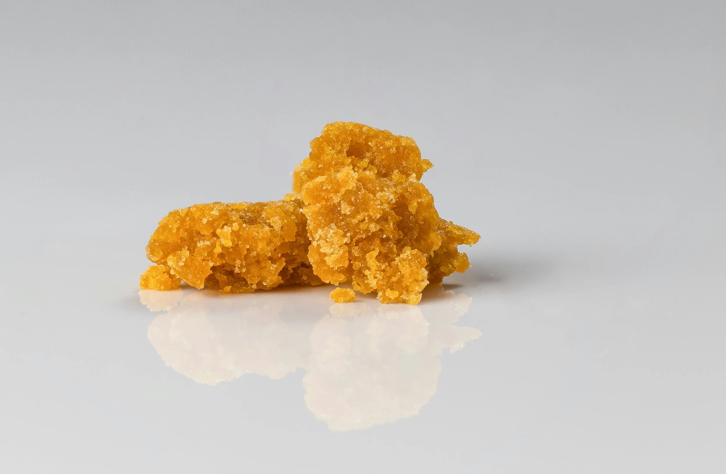 A small mound of golden-yellow cannabis wax concentrate with a crumbly texture, isolated on a white reflective surface.