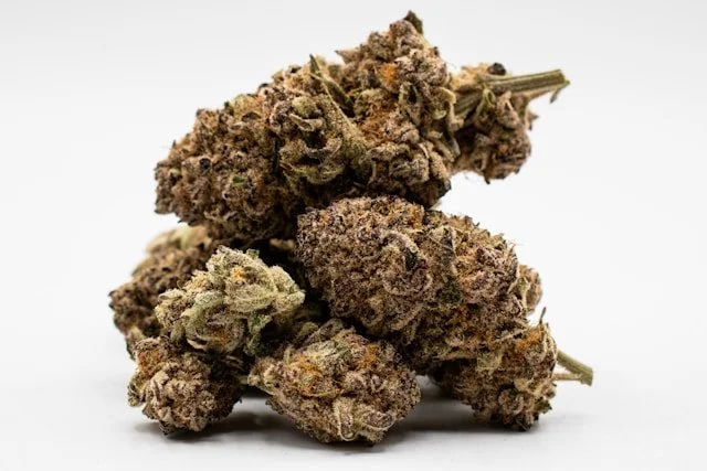 A close-up stack of premium dried cannabis flower buds showing various shades of green, purple, and orange hairs against a white background.