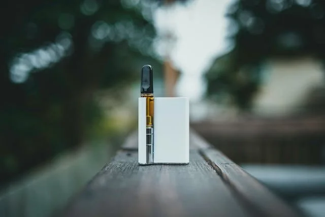 A sleek, modern cannabis vape cartridge and battery standing upright on a wooden railing with a soft-focus outdoor background.