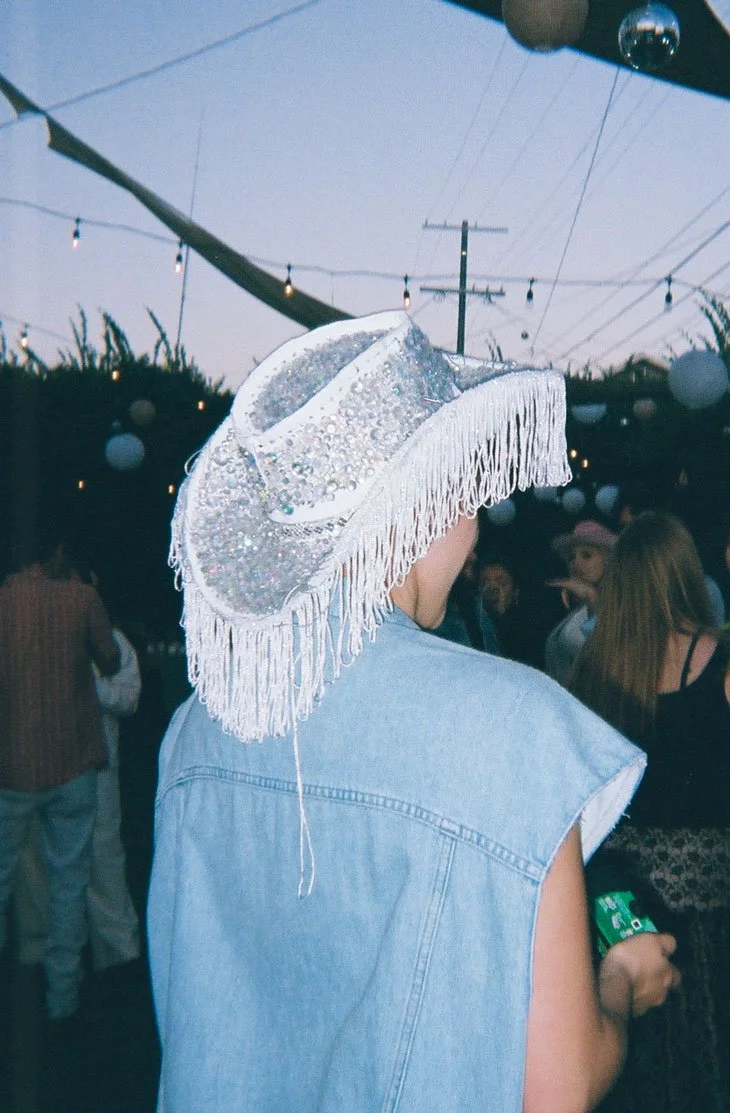 Person wearing a sequin cowboy hat with fringe and a denim jacket at an outdoor event.