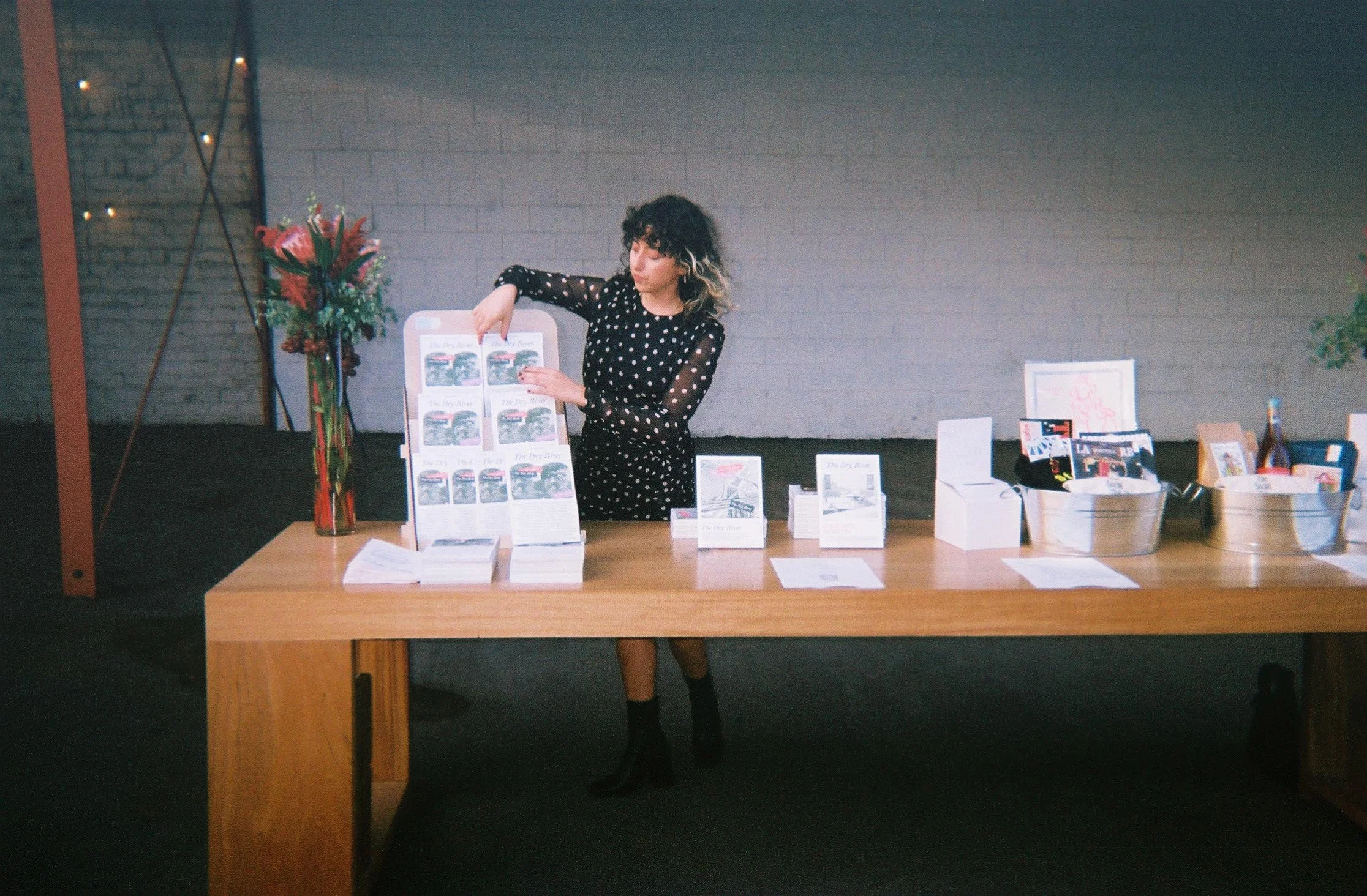 Person organizing books and magazines on a wooden table at an event with gift baskets and a flower vase.