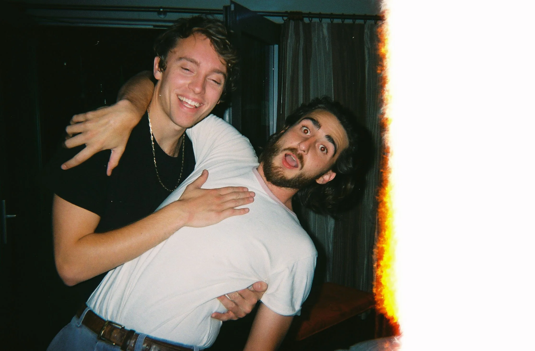 Two people laughing and embracing, with one wearing a black shirt and chain necklace, and the other in a white shirt, inside a room with curtains in the background.