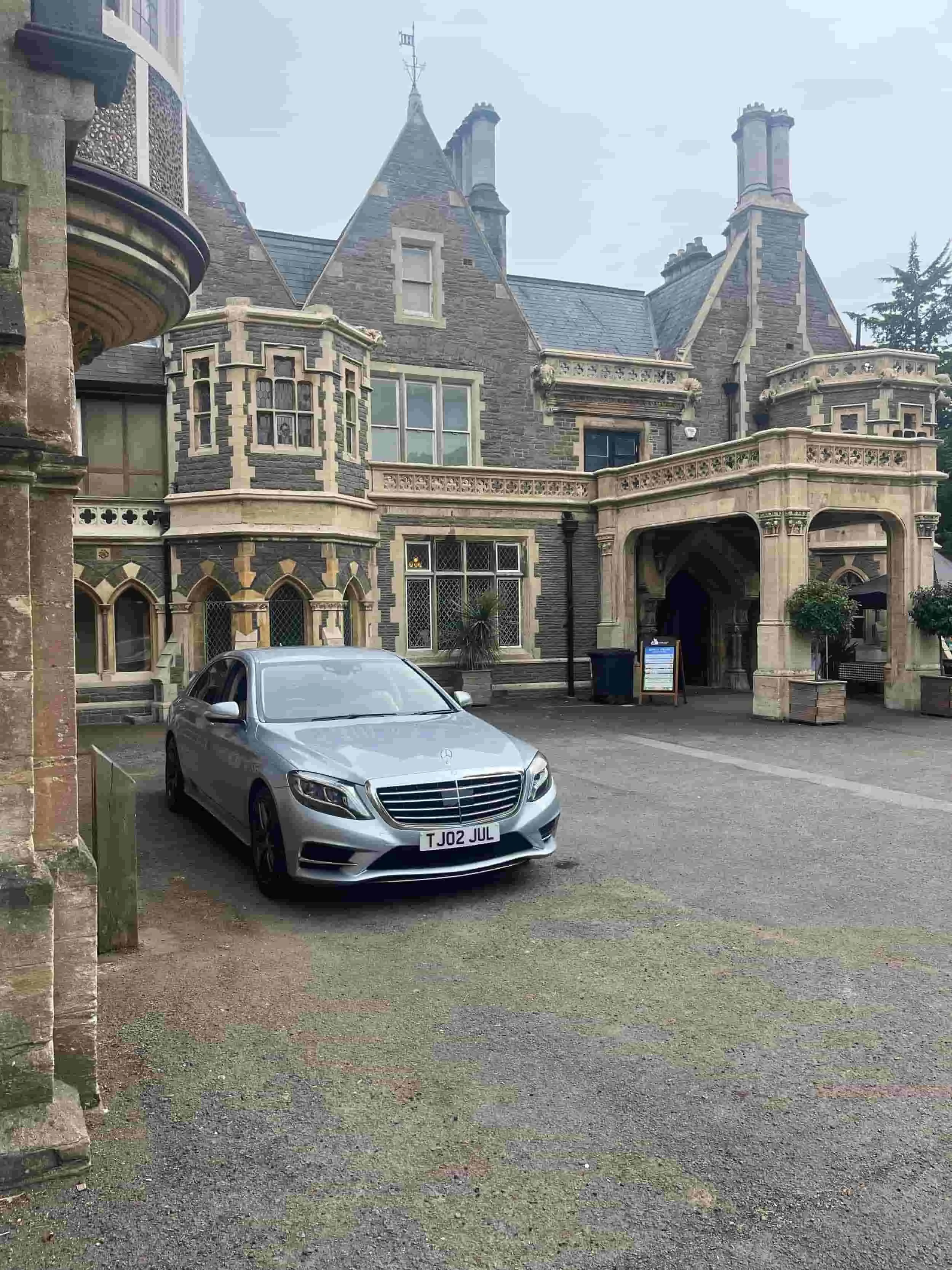 A silver Mercedes-Benz with a chauffeur waits outside a large, ornate stone mansion with arched doorways and tall chimneys.