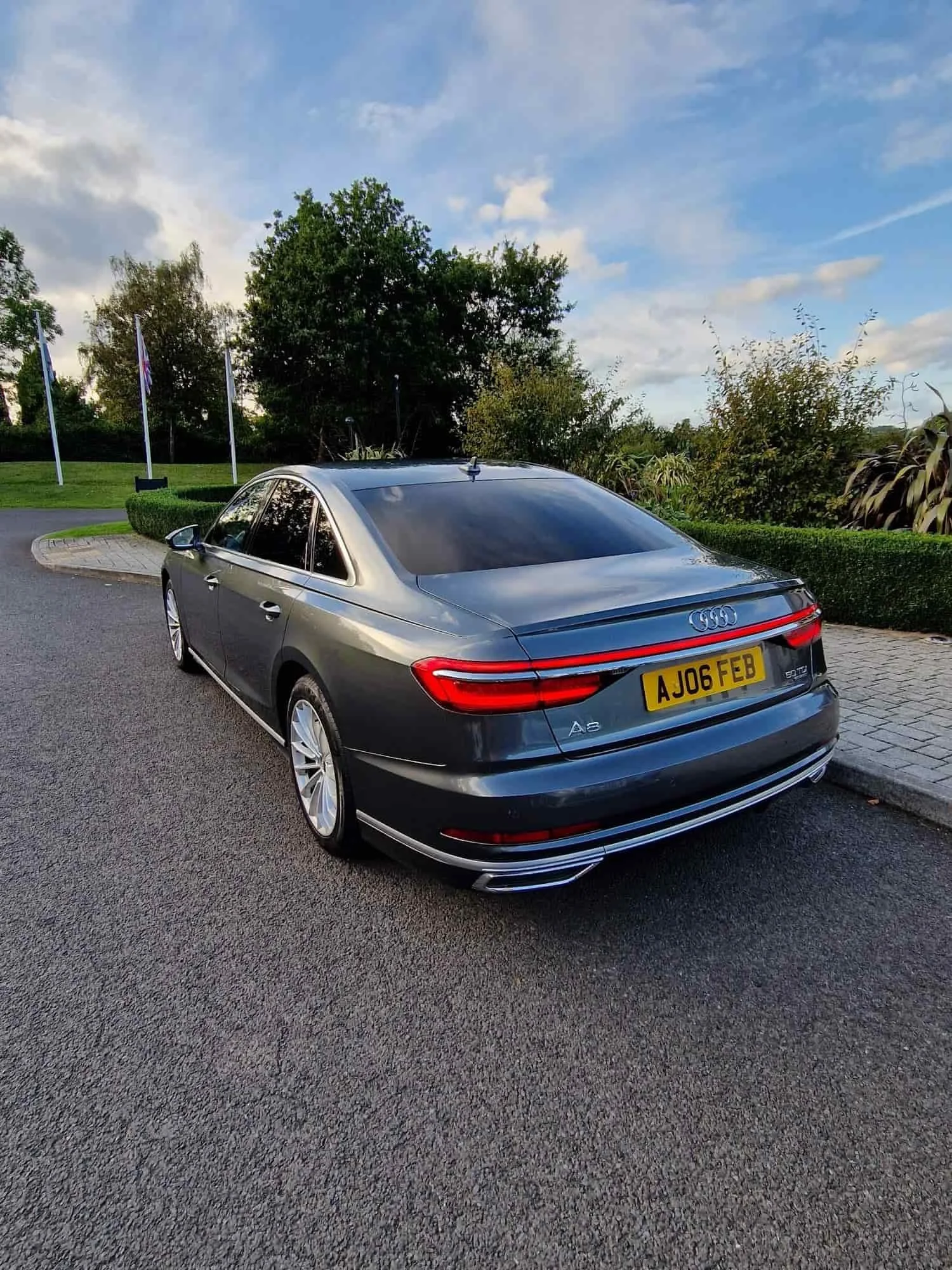 A grey Audi A8 sedan parked on a road near greenery, viewed from the rear left side, under a partly cloudy sky.
