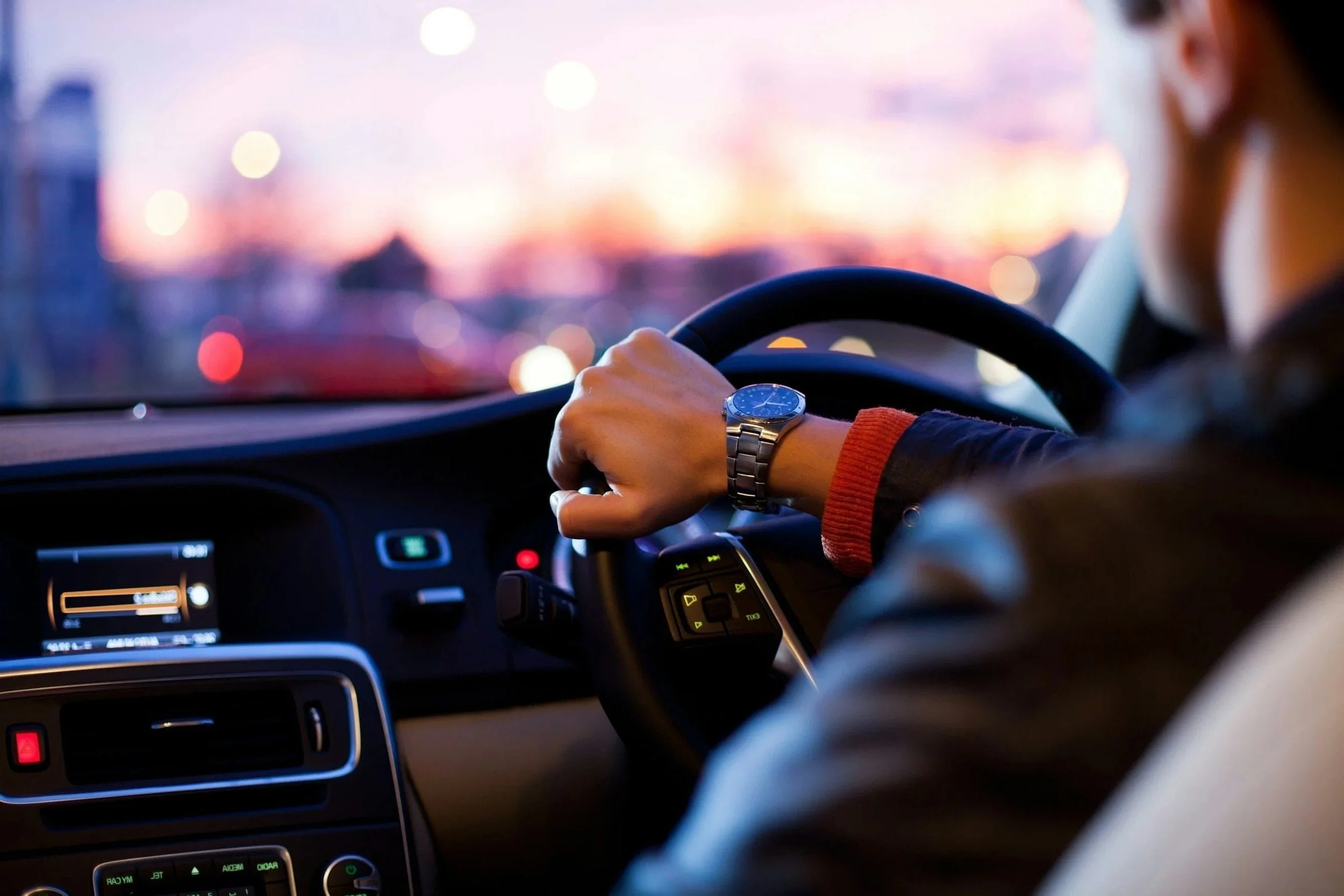 A person driving a car with a watch on their left wrist, on a city street at dusk with blurred city lights visible through the windshield.