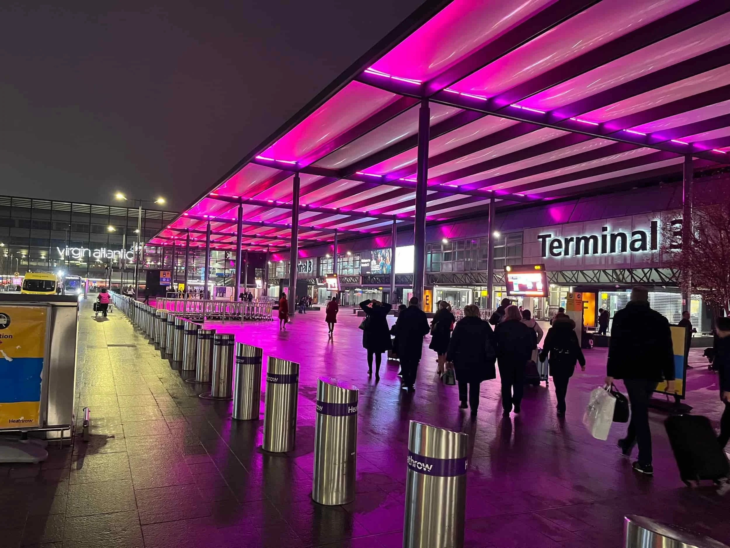 People walk outside Heathrow Terminal 3 at night under a canopy lit with bright pink and purple lights.