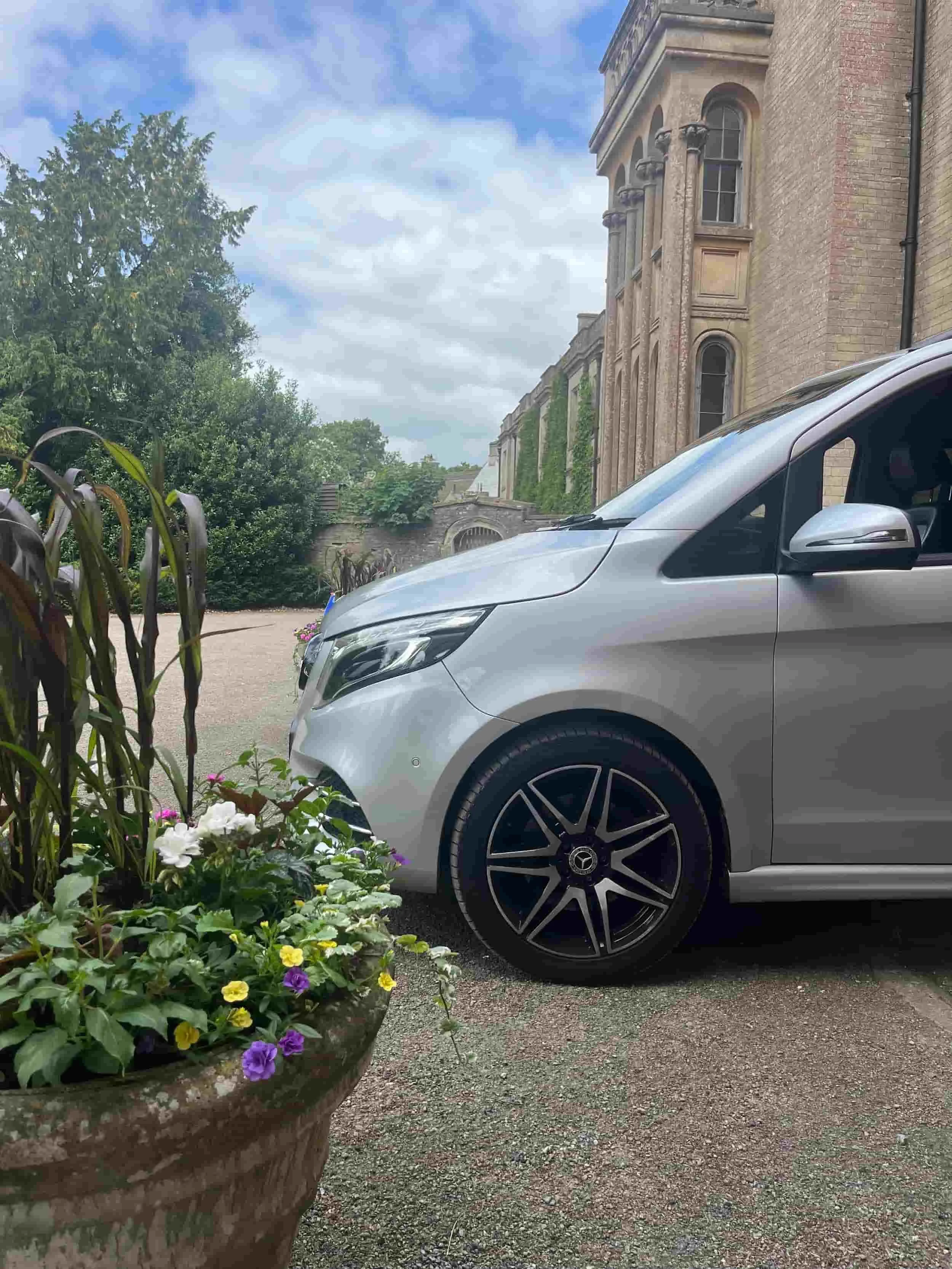 A silver car parked near a historic building, with a flower planter in the foreground and greenery in the background.