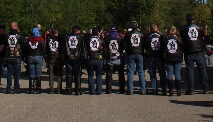 Group of people standing together outdoors, wearing matching black leather vests with a logo on the back.