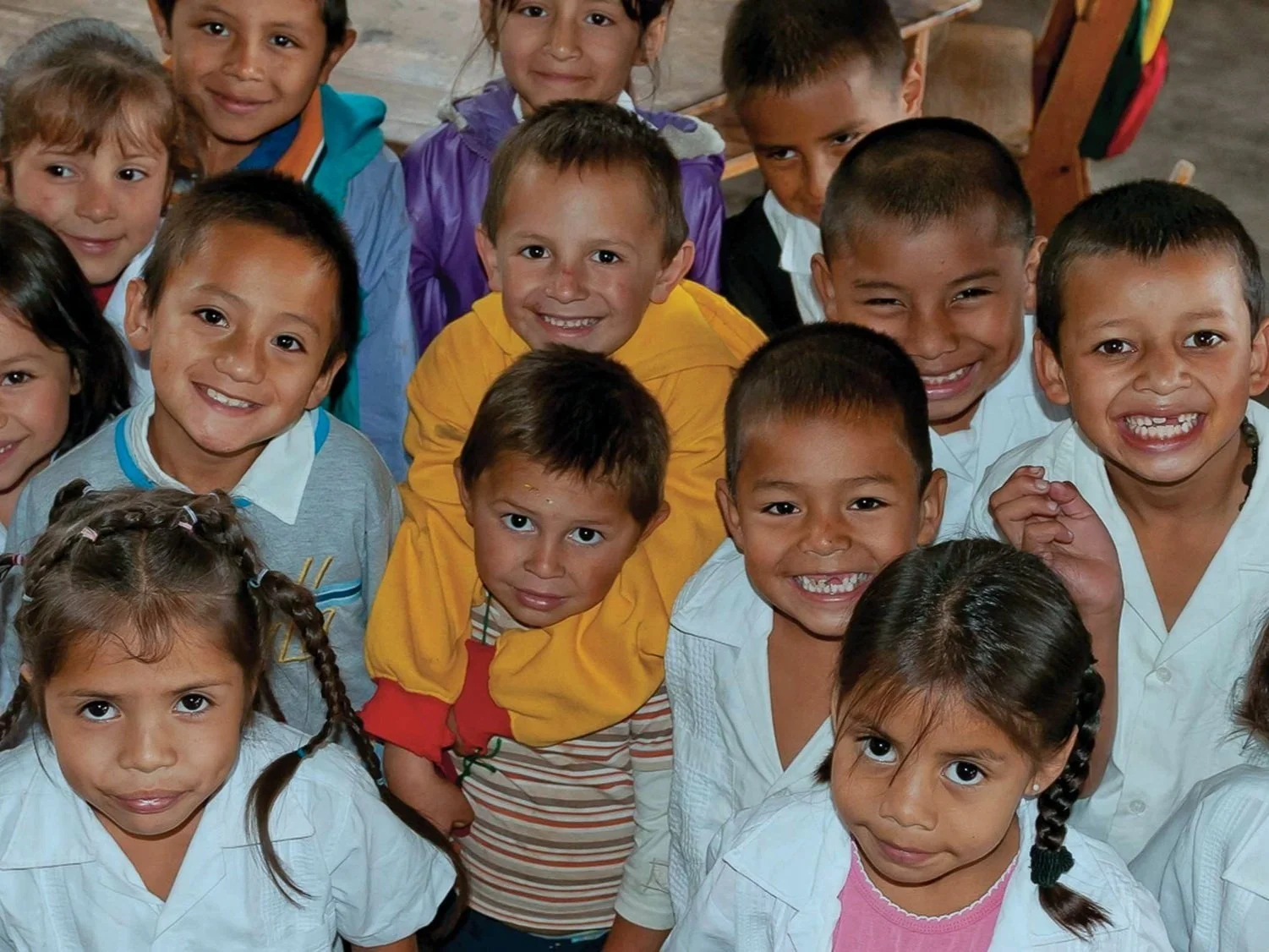 Group of smiling children in classroom, some wearing white uniforms, others in colorful clothing, looking at the camera.