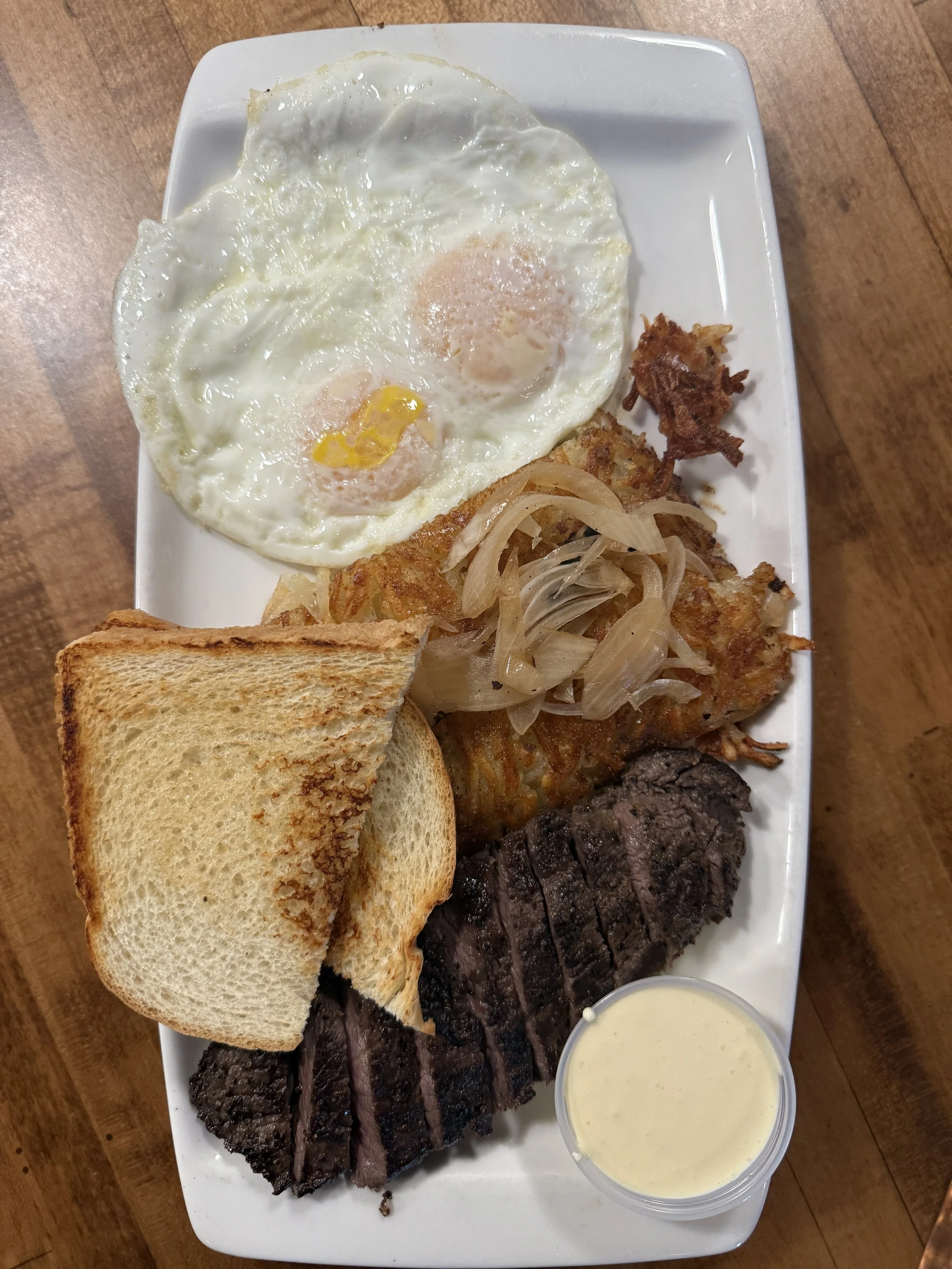 A breakfast plate with two fried eggs, toast, grilled steak, sautéed onions, and a side of creamy white sauce or dressing.