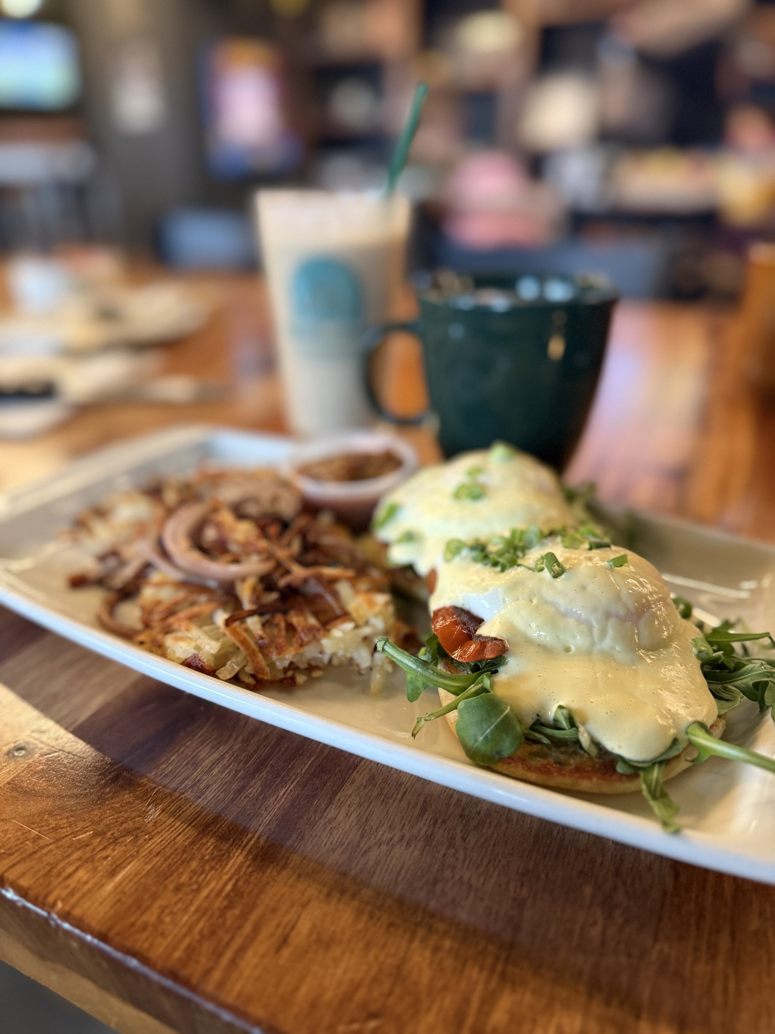 Plate with eggs benedict topped with hollandaise sauce and green onions, hash browns with onions, and a side of salsa, served with a coffee mug and a smoothie in a restaurant setting.