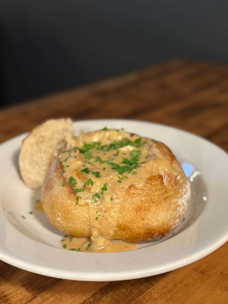 A baked bread bowl filled with creamy seafood sauce, garnished with chopped parsley, on a white plate with a piece of bread on the side.